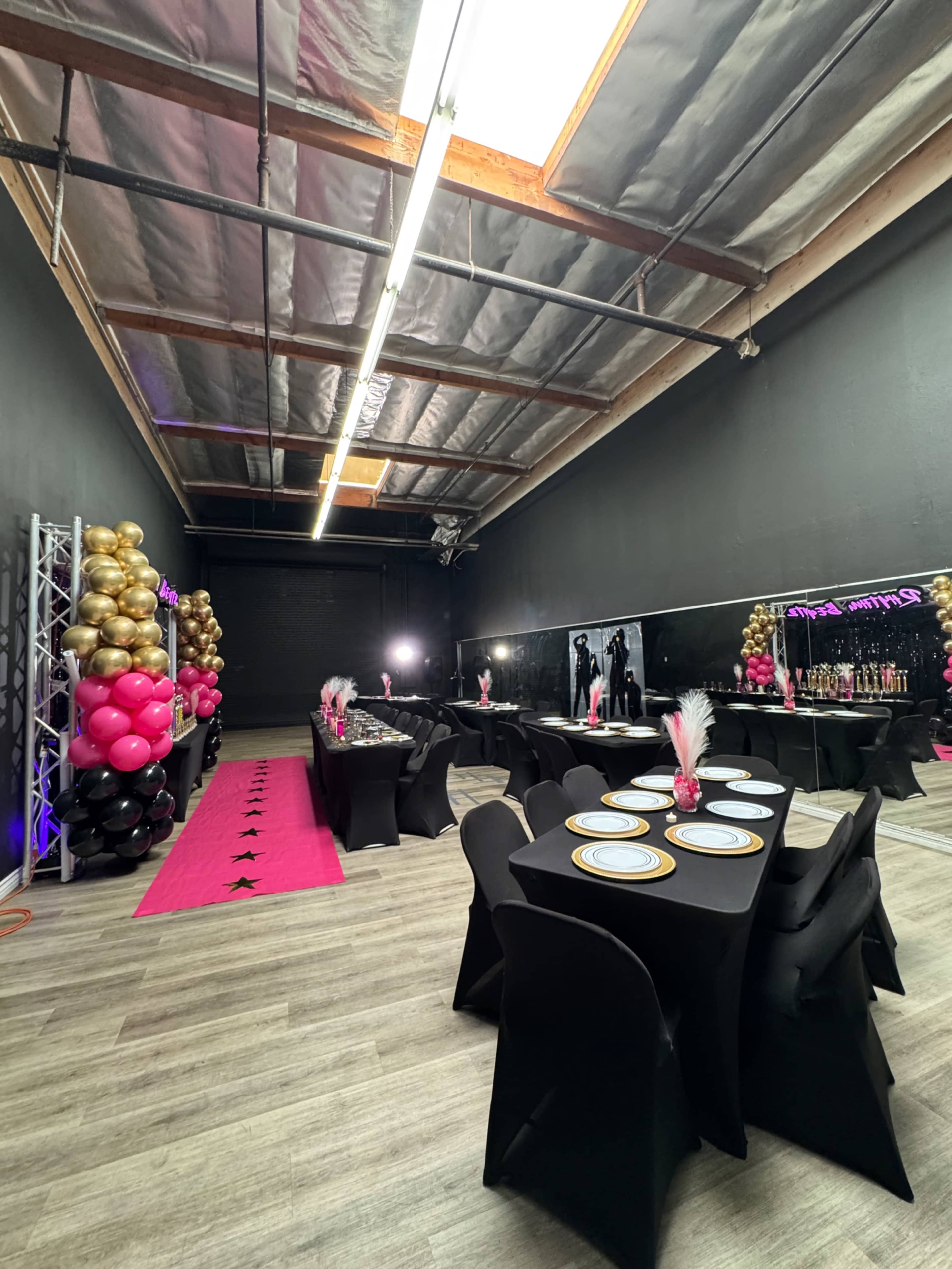 The image shows a party setup in a spacious room with black walls, adorned with black tables draped in black cloth, set with plates and pink and gold balloon decorations.