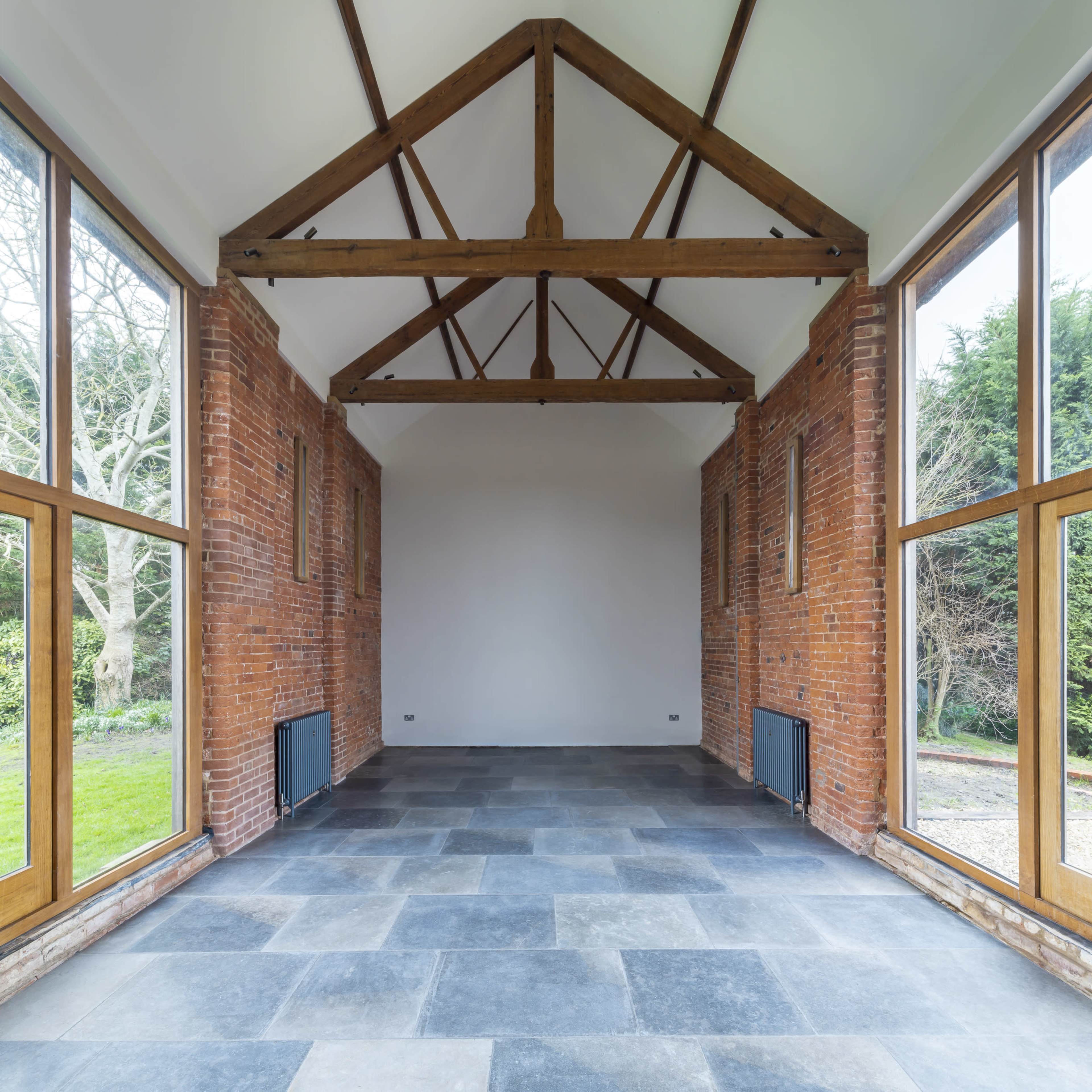 The image shows a spacious indoor room with exposed wooden beams, brick walls, large windows on either side, and a slate-tiled floor.