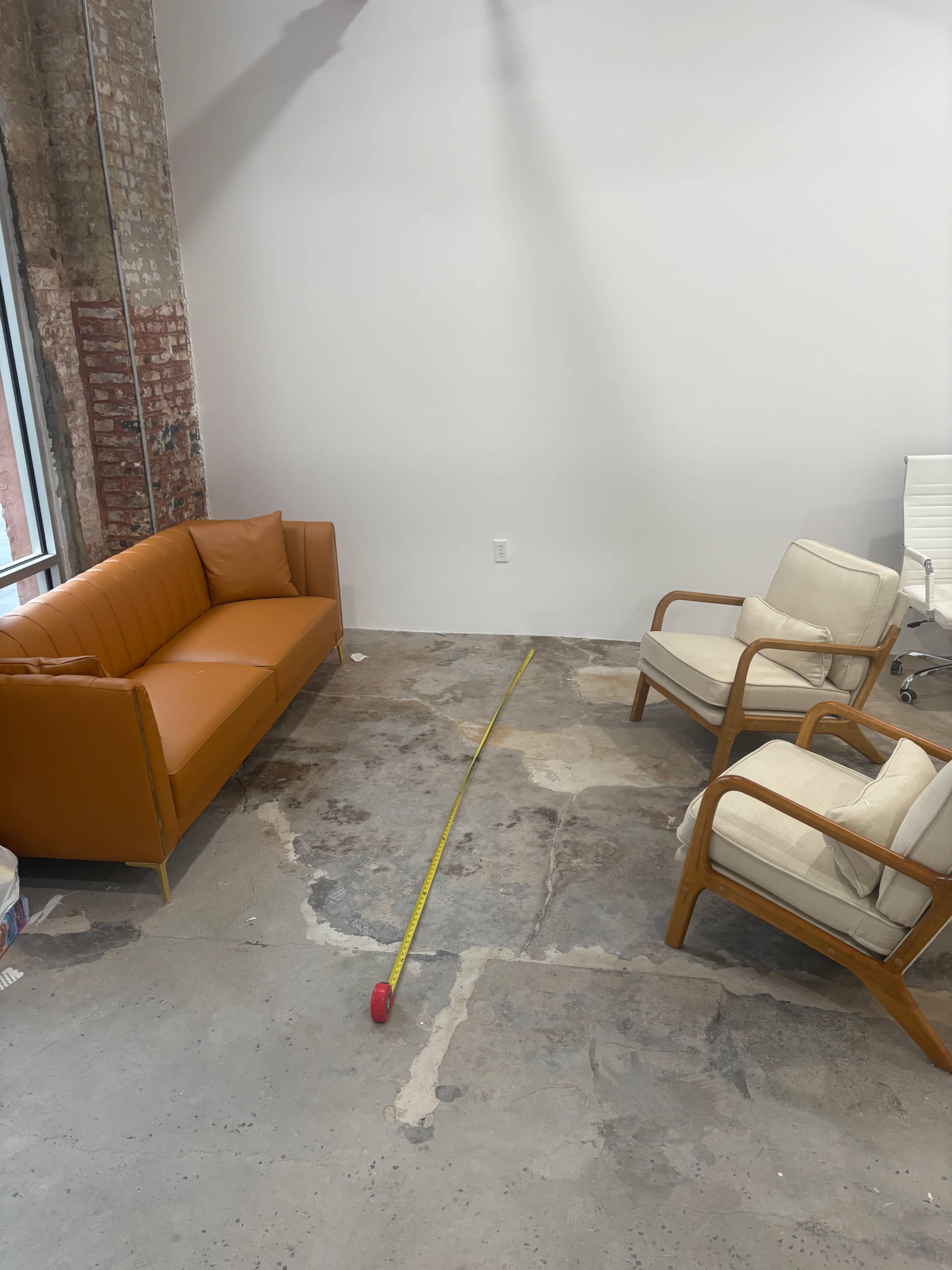 The image shows a room with an orange couch and two cream-colored armchairs positioned on a concrete floor, with a yellow measuring tape running across the floor.