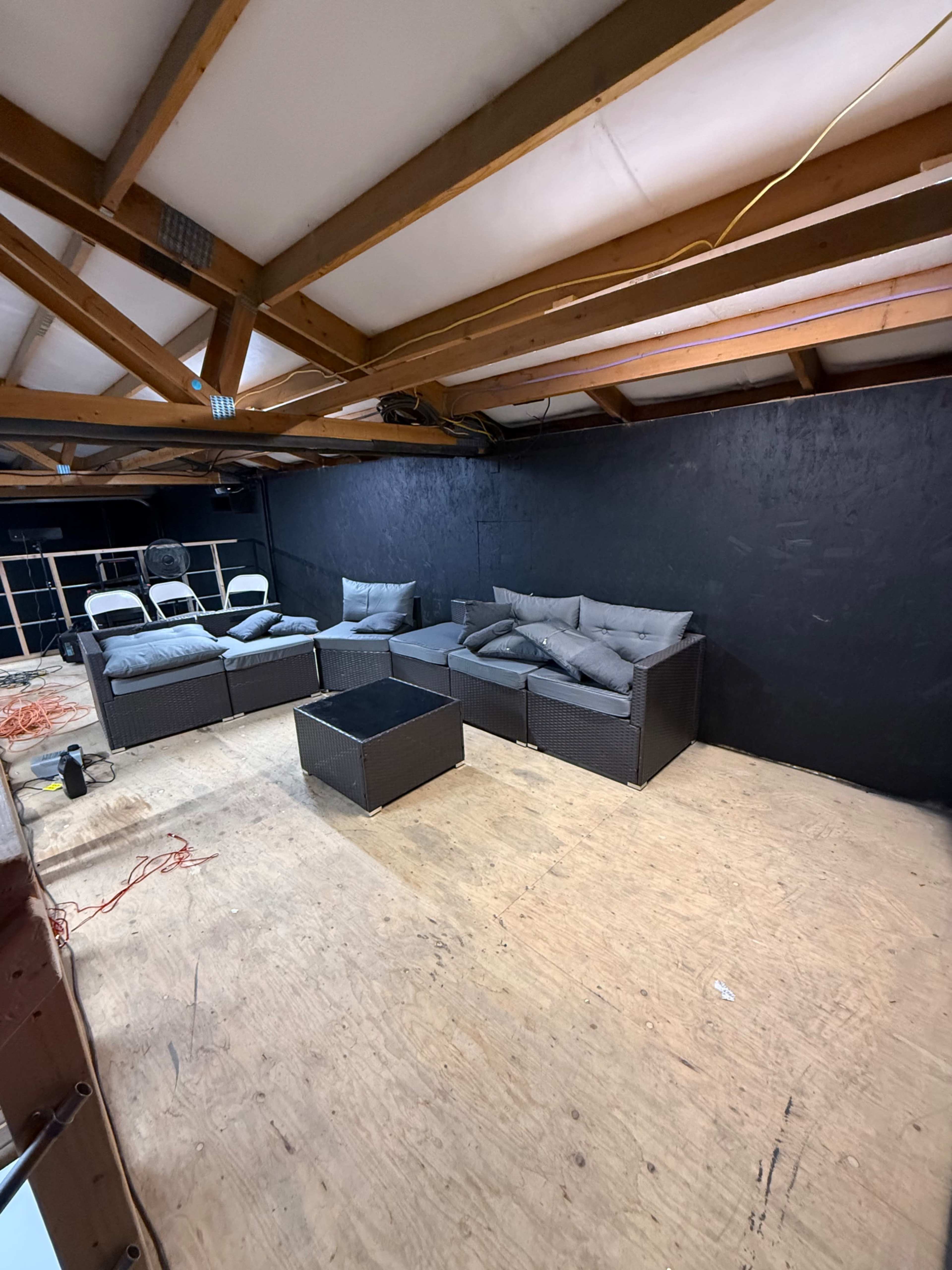 The image shows a seating area with dark wicker furniture and cushions arranged on a wooden floor in a room with exposed beams and a black wall.