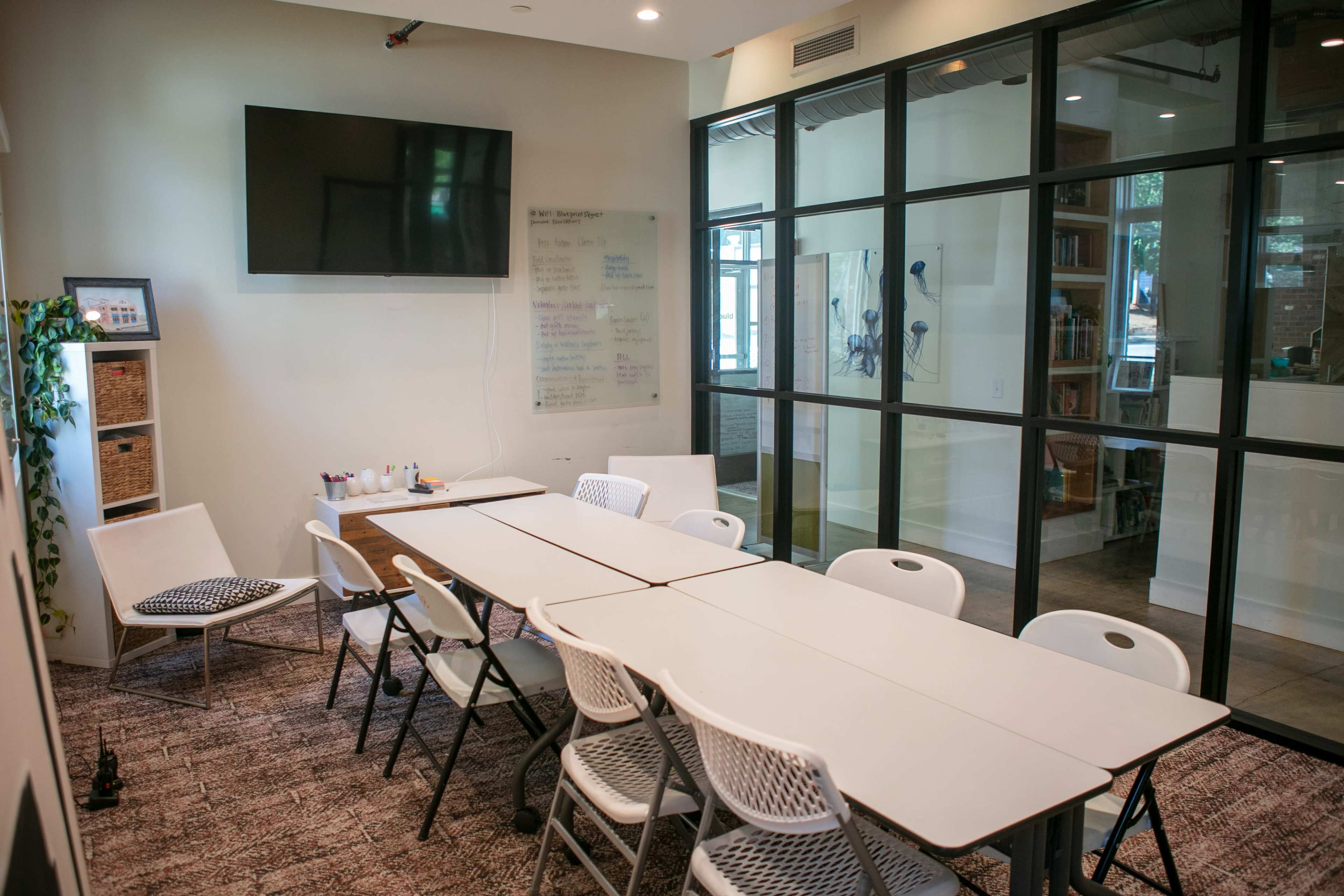 A conference room features a long table surrounded by white chairs, a wall-mounted TV, and a whiteboard with notes, with a glass partition leading to a bookshelf area.