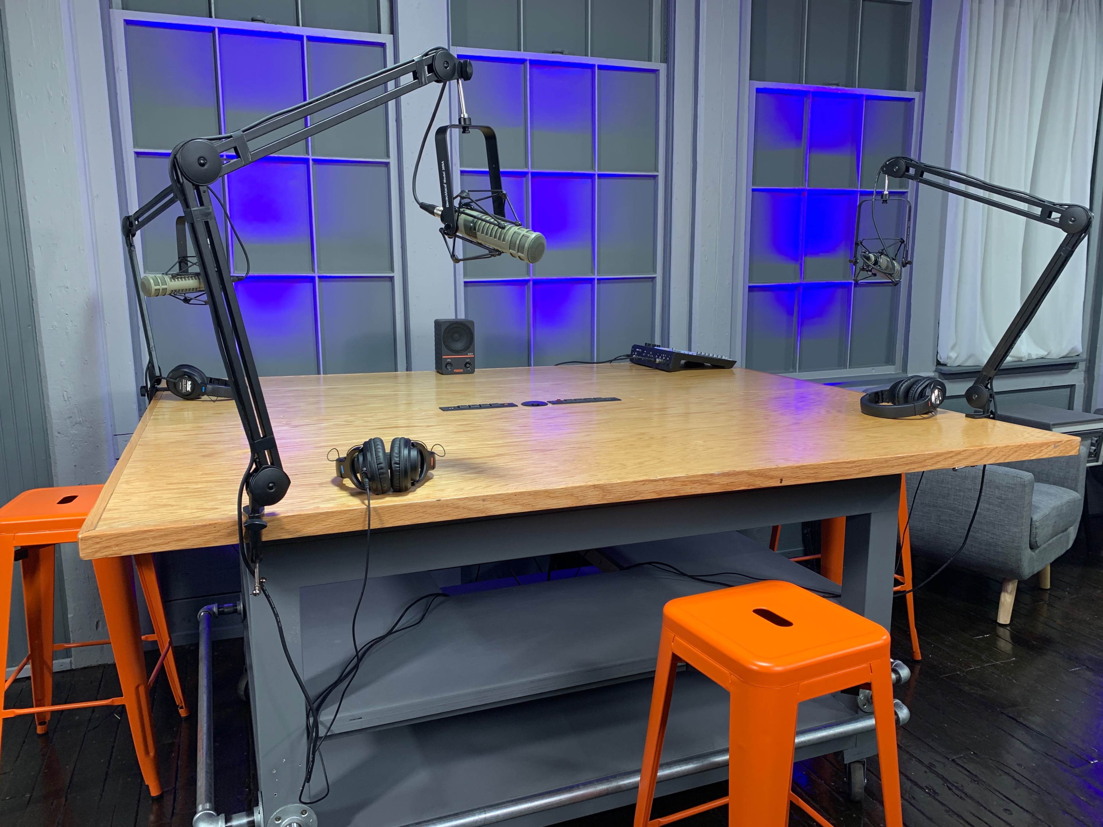 The image shows a podcast recording studio featuring a wooden table with microphone stands, headphones, and orange stools.