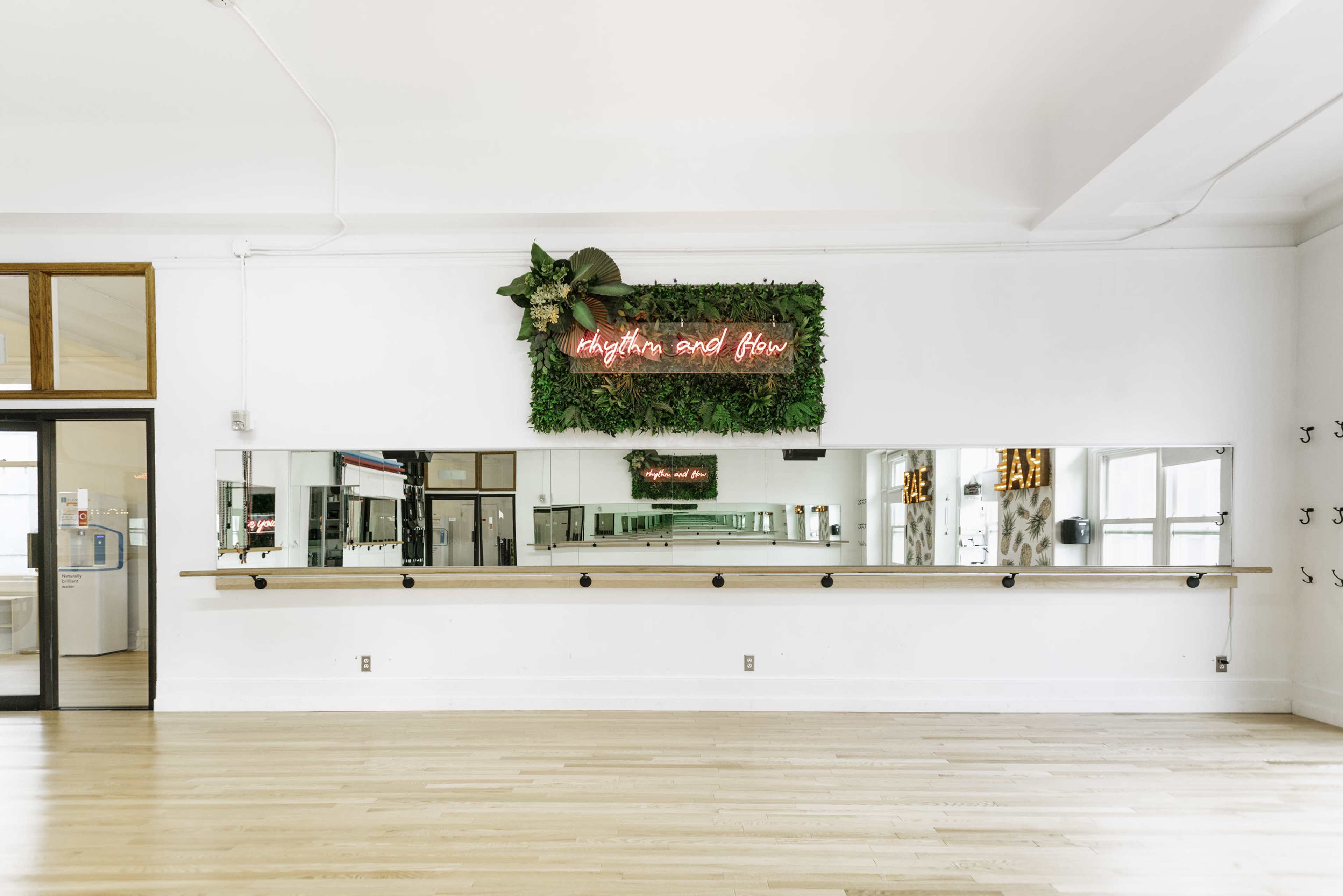 A large, well-lit room features a wall-mounted mirror and a wooden barre beneath a neon sign that reads "rhythm and flow," surrounded by greenery.