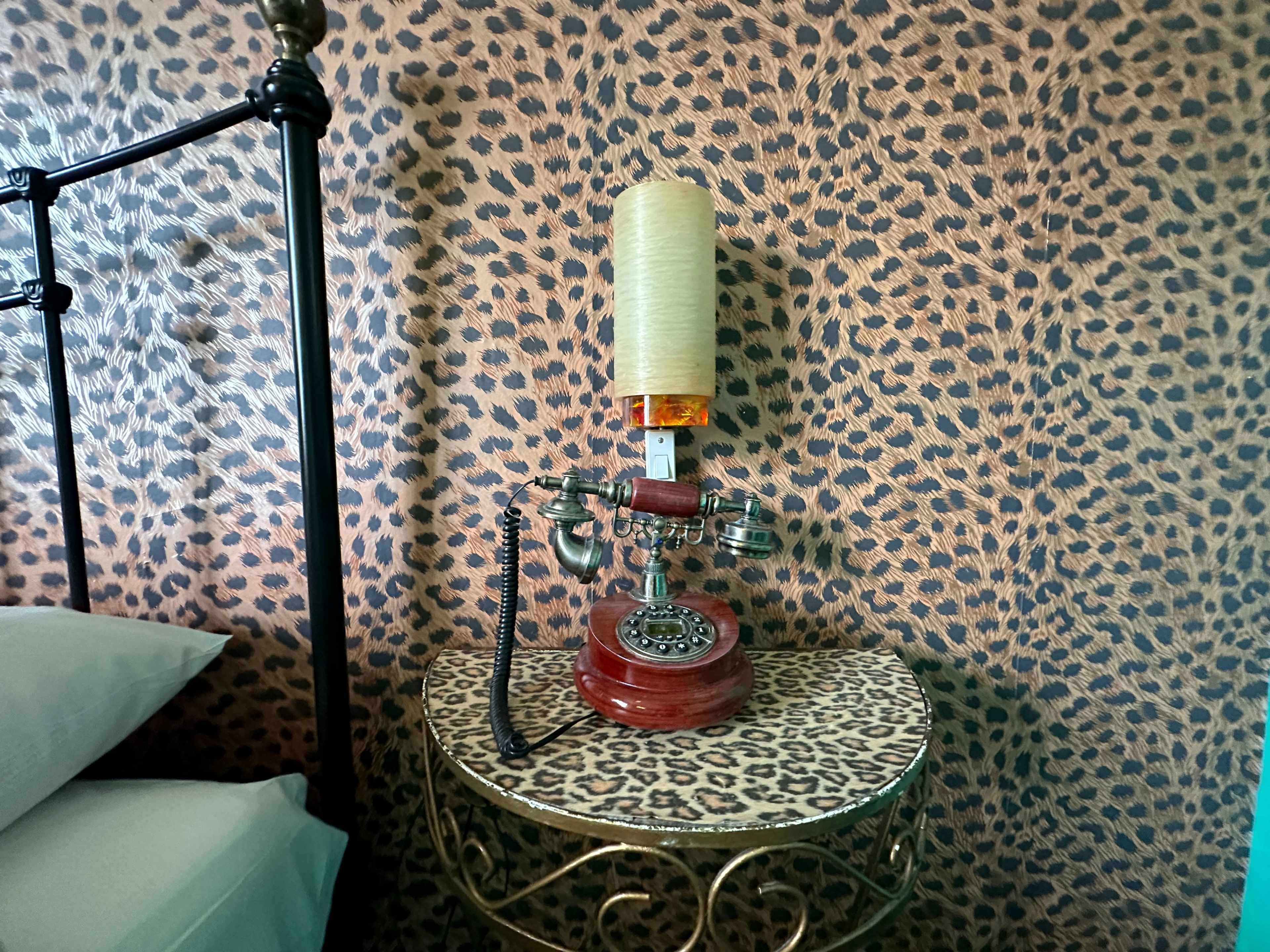 An old-fashioned rotary telephone is placed on a leopard-print side table next to a bed, against a matching leopard-print wallpaper.