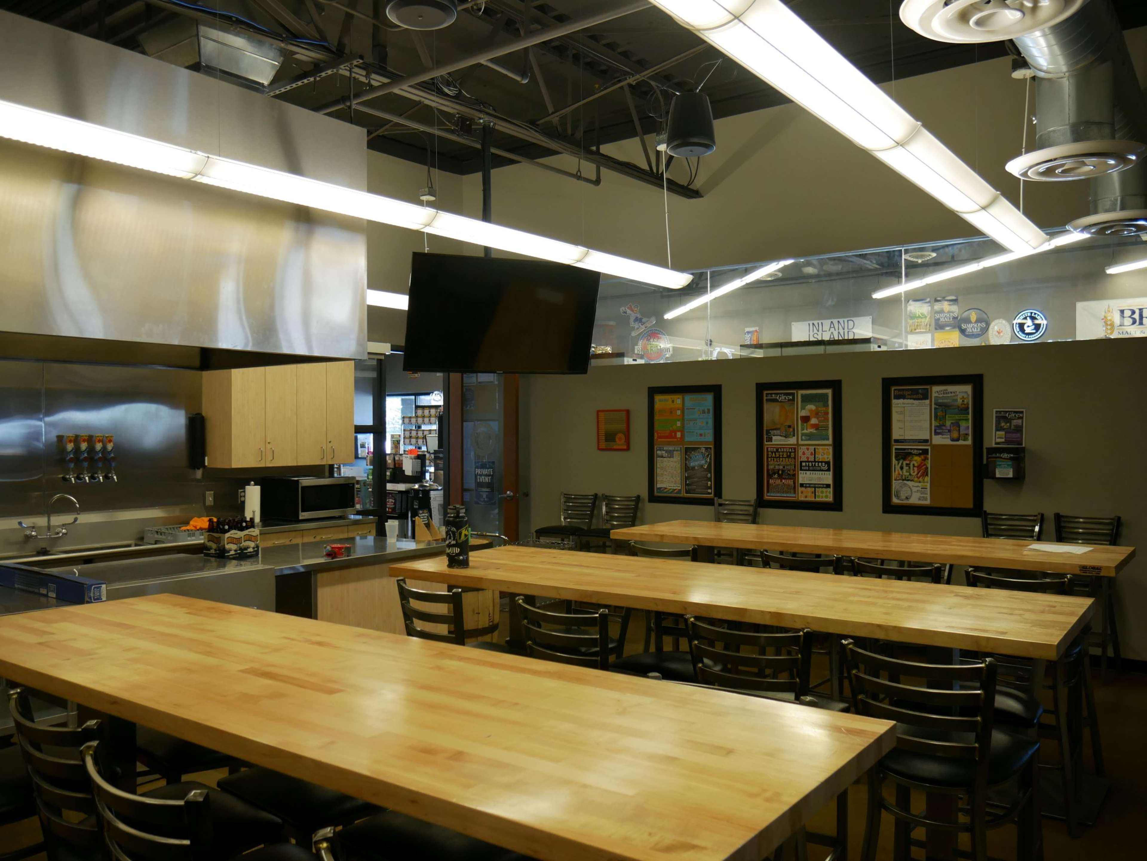 A spacious kitchen and dining area with stainless steel appliances, long wooden tables, and a large screen mounted on the wall.