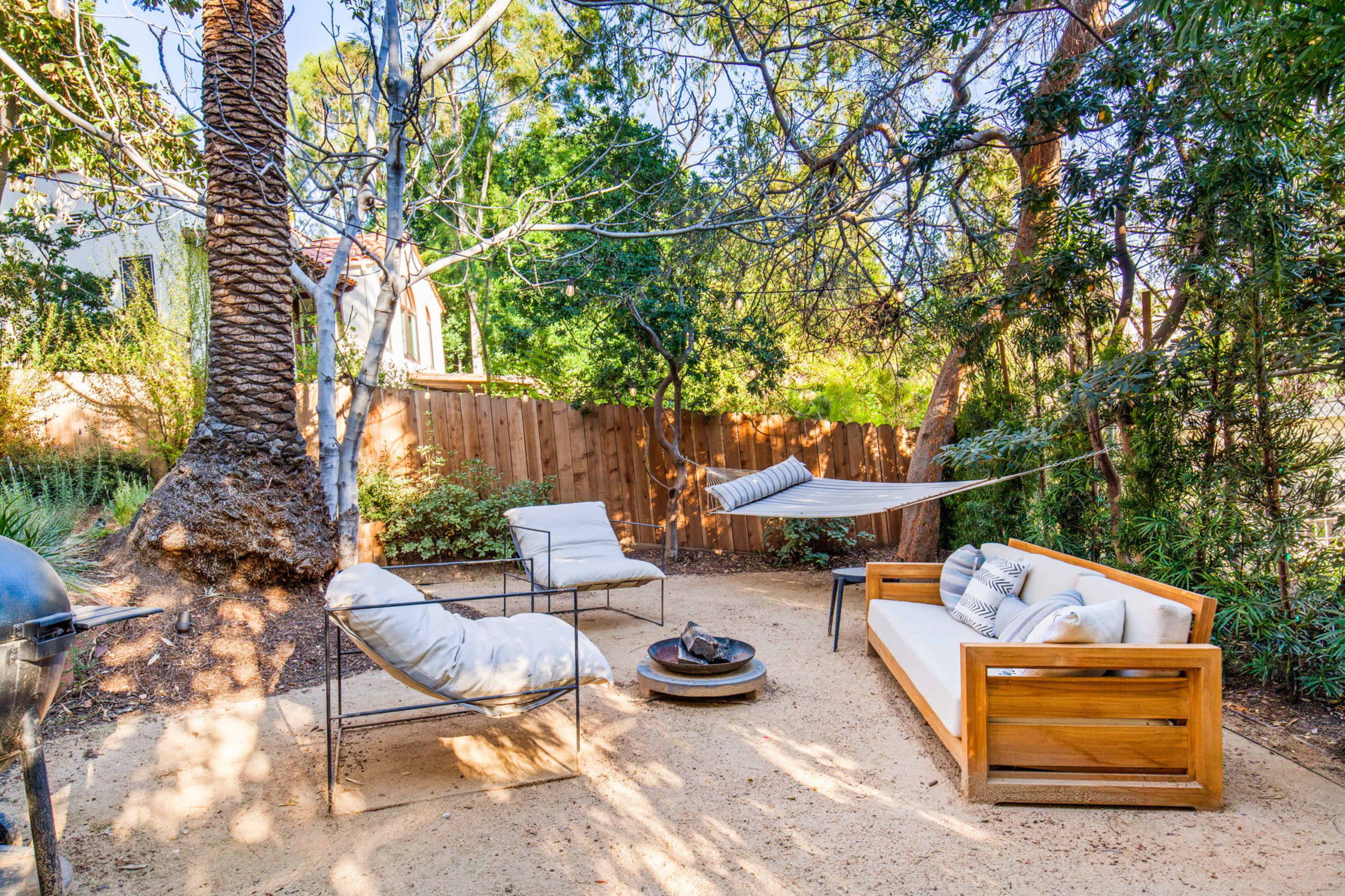 A backyard seating area with a wooden couch, two chairs, a fire pit, and a hammock suspended between trees.
