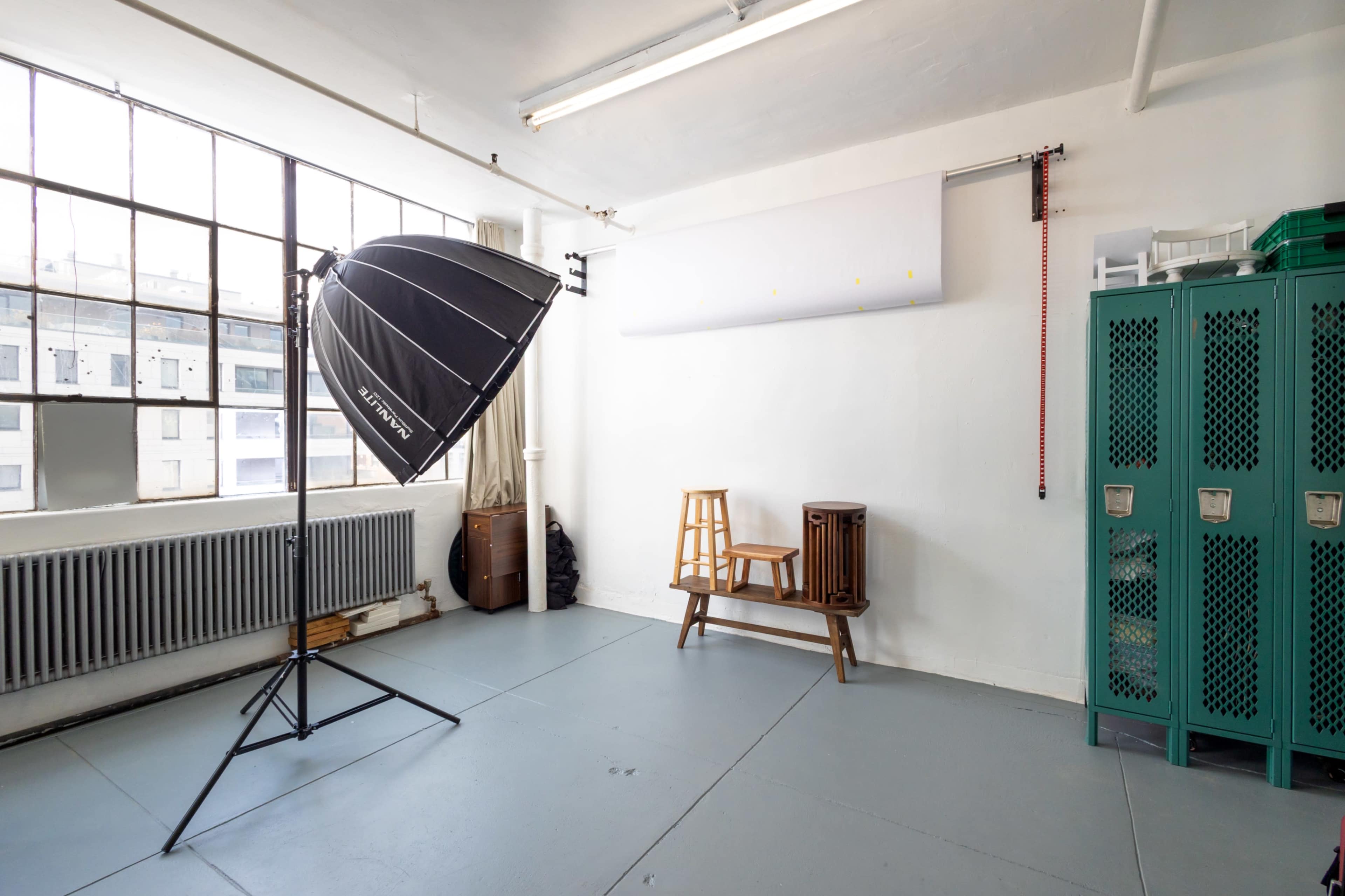 The image shows a photo studio with a large light umbrella set up next to a wooden stool and a bench against a white wall, with green lockers in the corner.