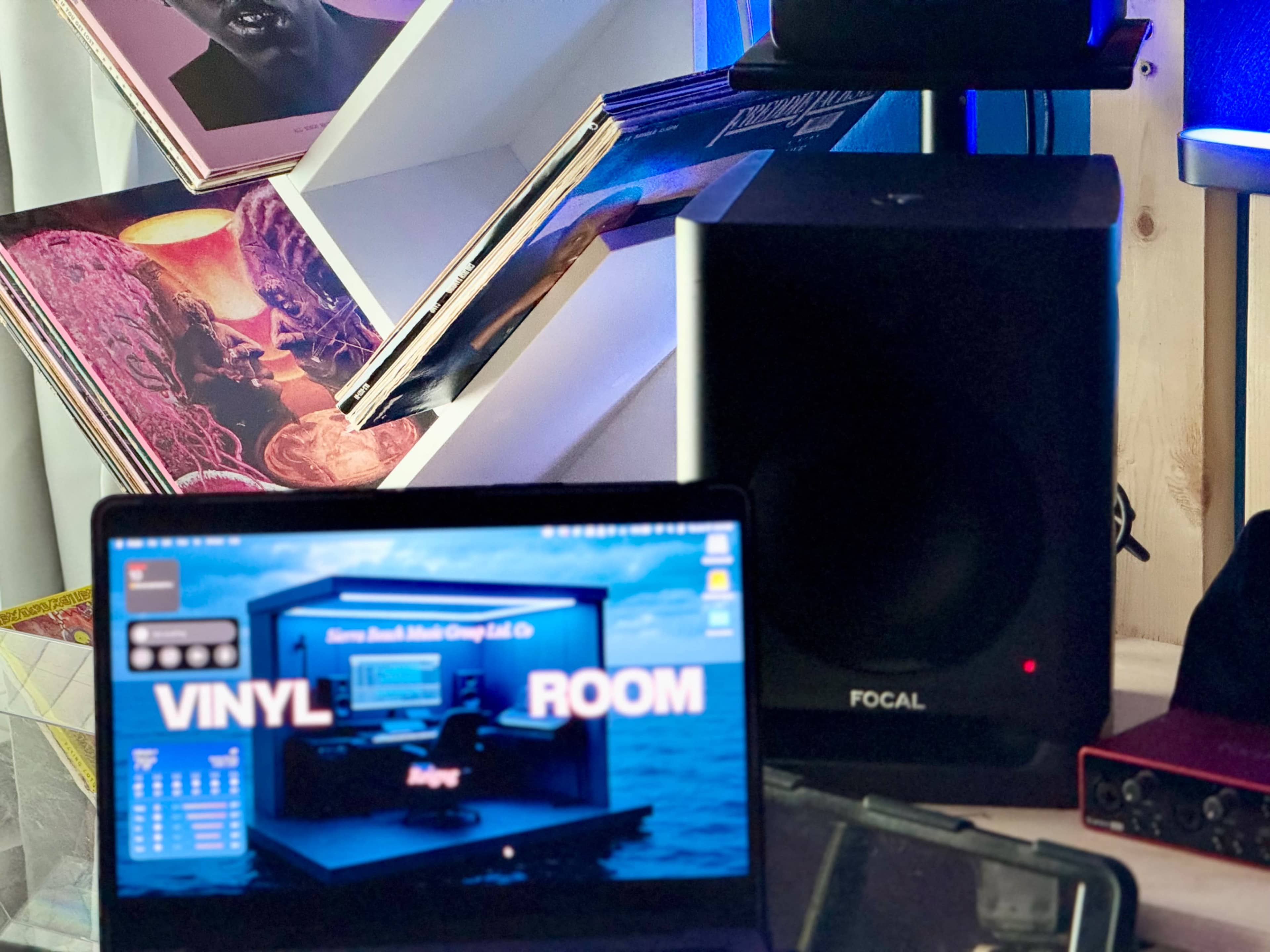 The image shows a laptop displaying a "Vinyl Room" interface next to a speaker and a shelf filled with vinyl records.