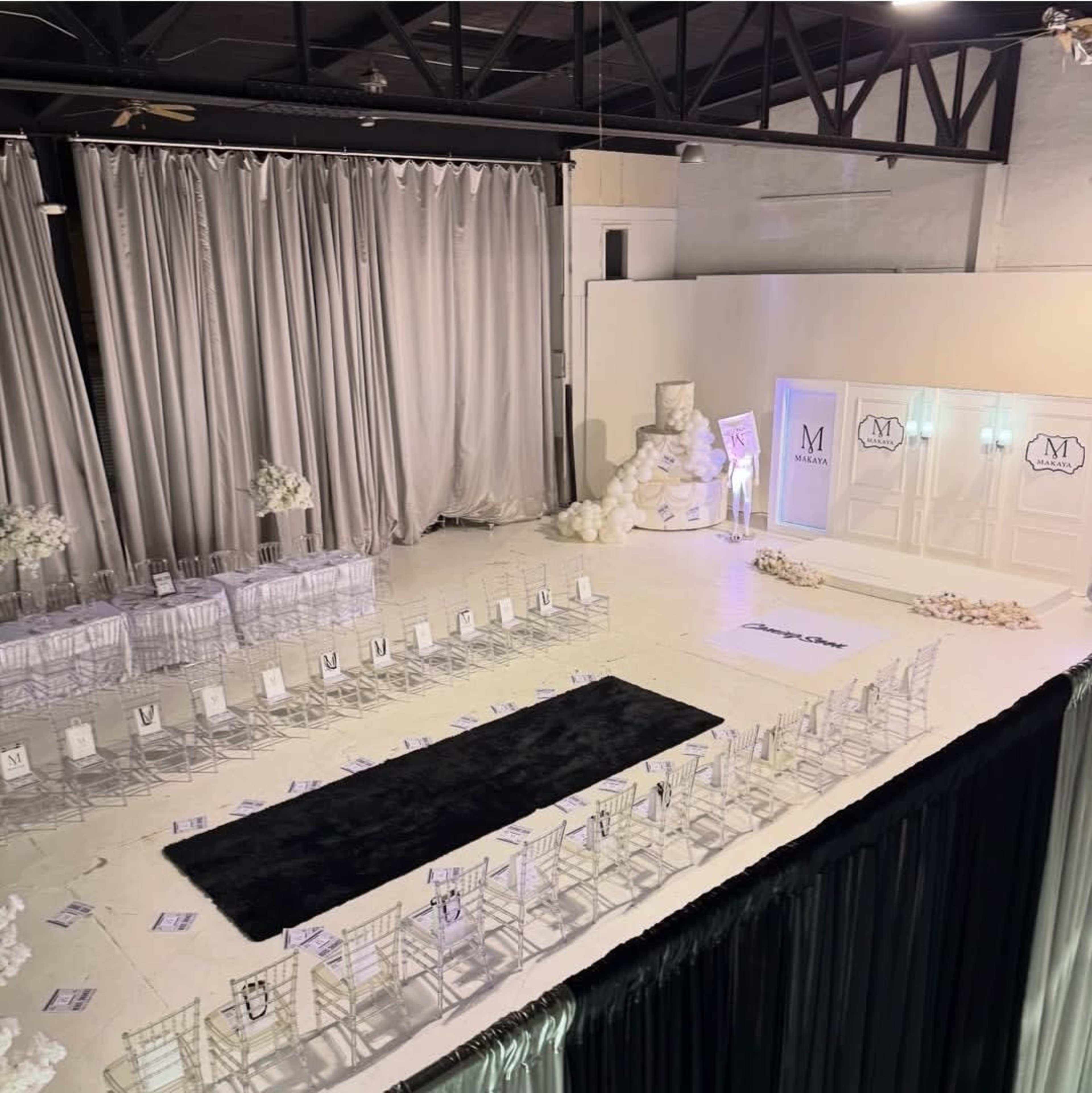 The image shows a spacious event venue set up for a wedding, featuring rows of chairs arranged facing a central aisle, adorned with white decorations and a large cake display.