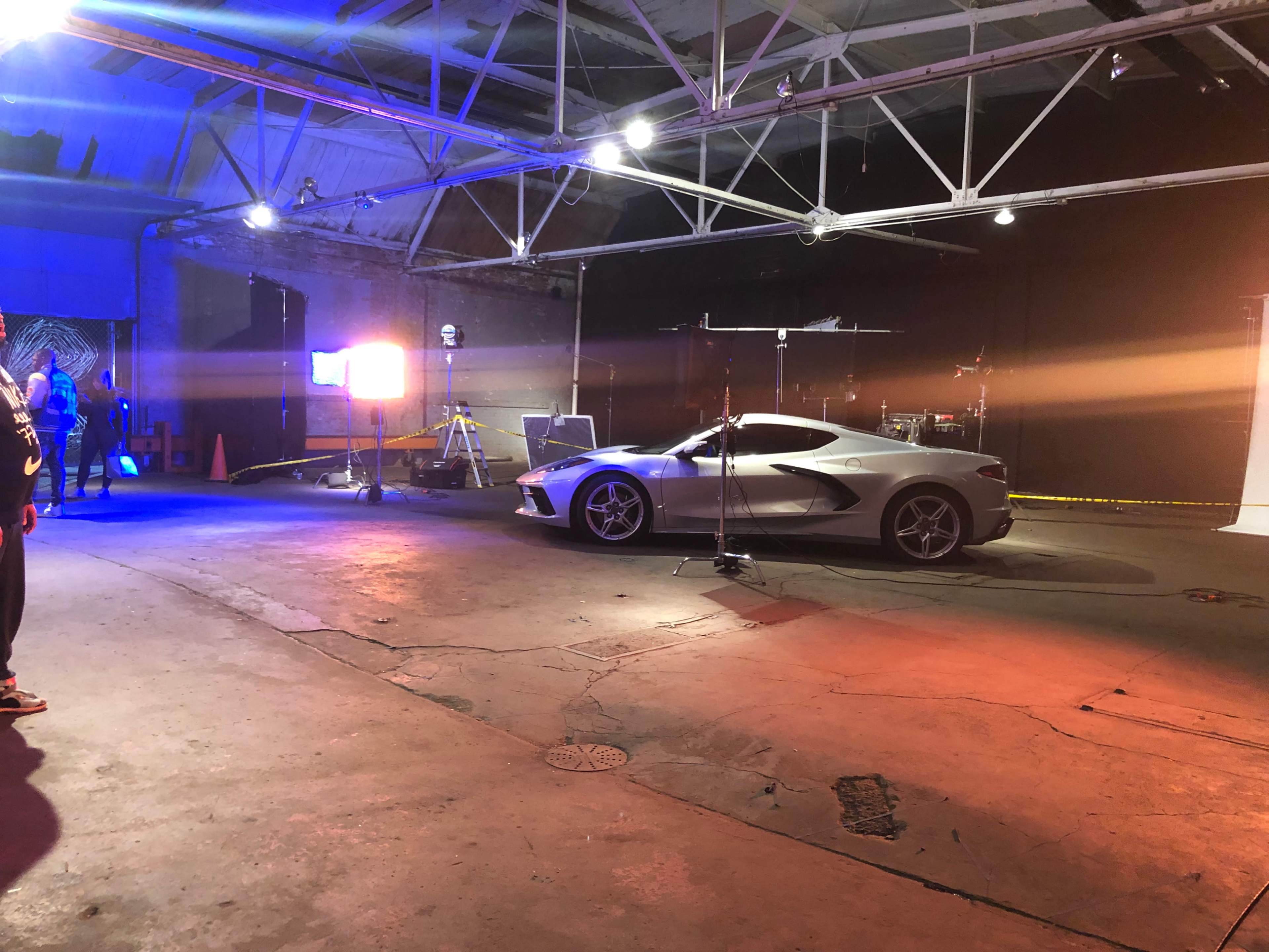 A sleek silver sports car is parked in a well-lit studio space with equipment and a drum set visible in the background.