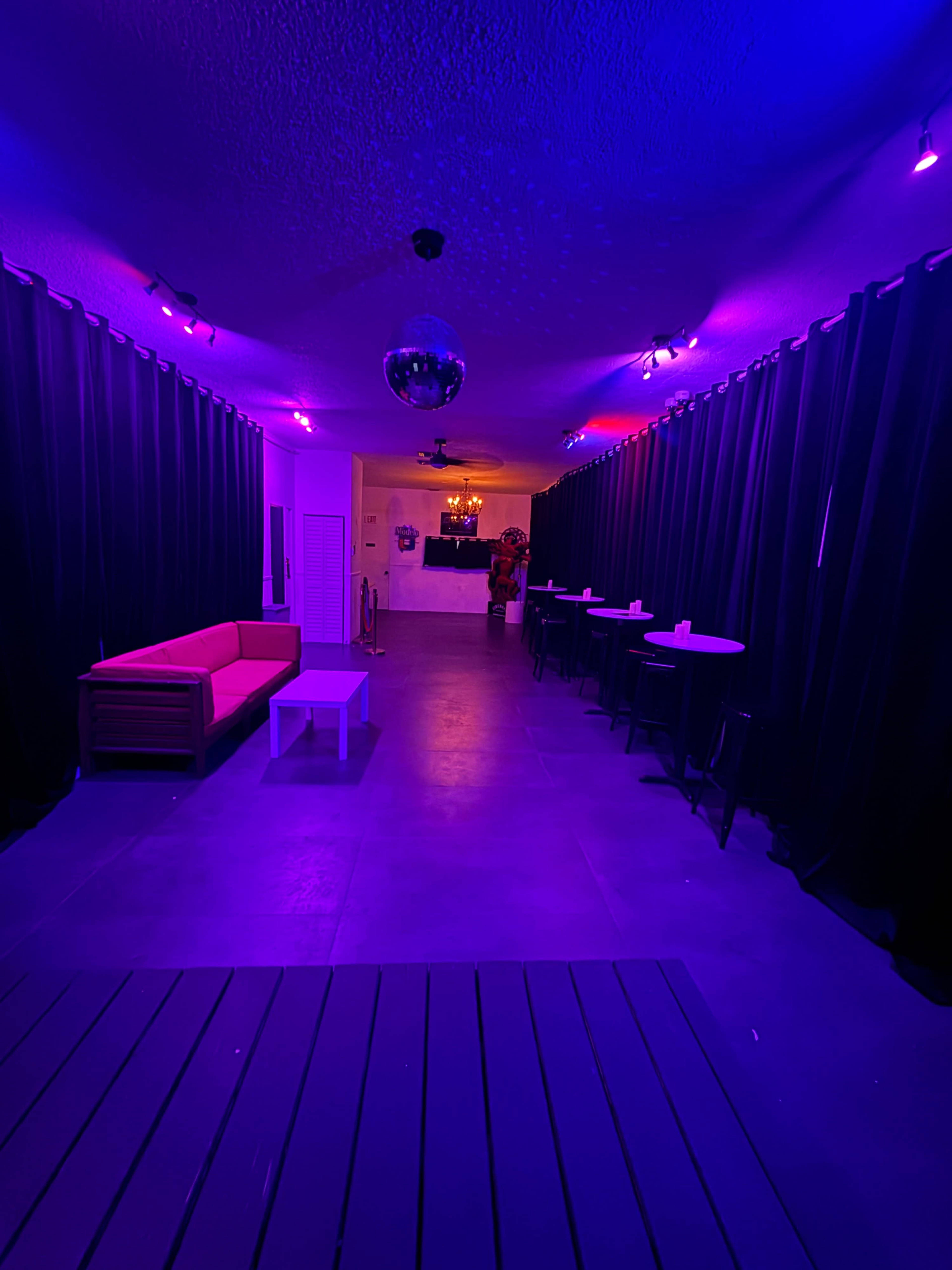 The image shows a dimly lit corridor with purple lighting, featuring a red sofa on one side and several high tables along the walls.