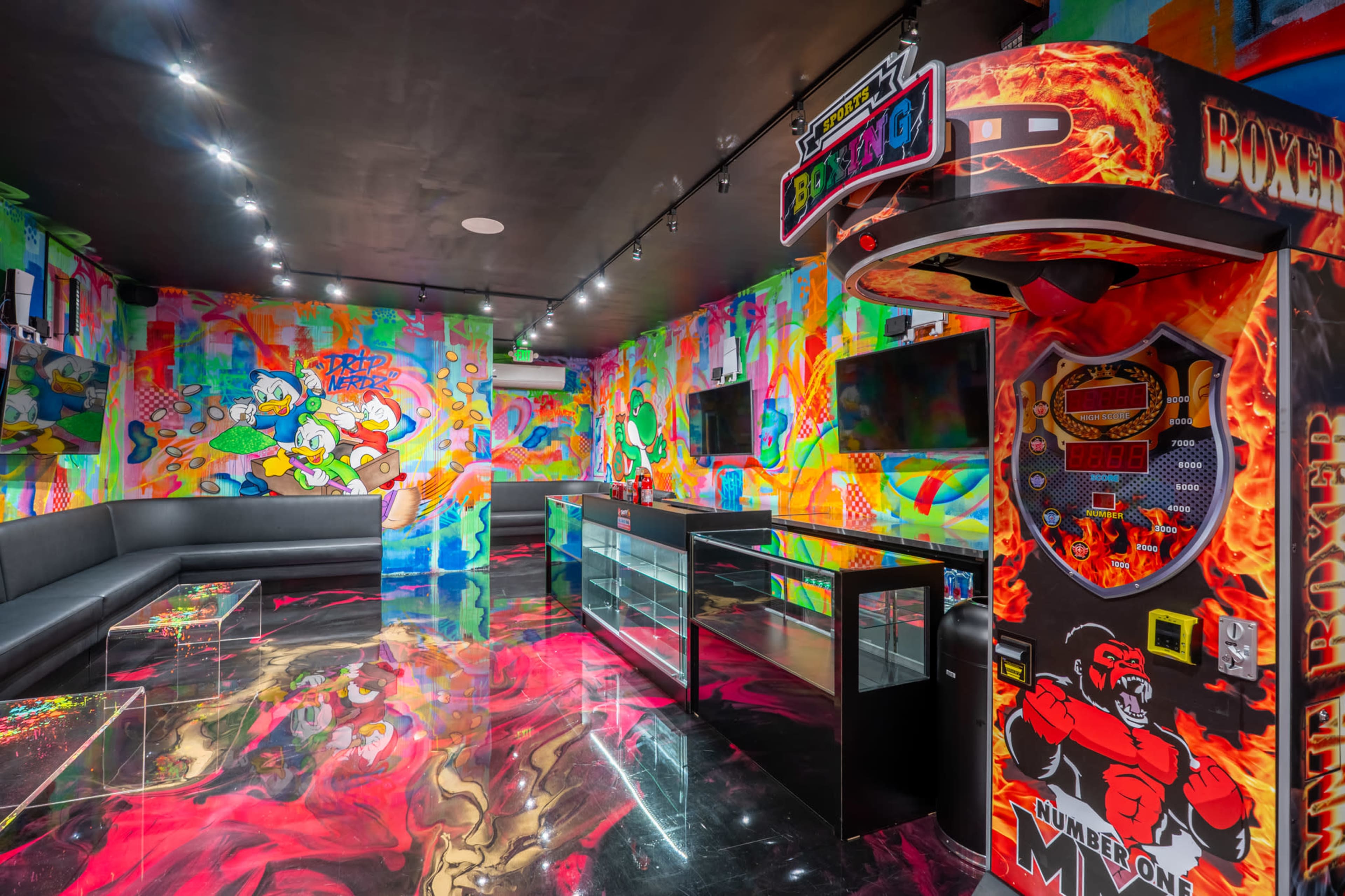 The image shows a vibrant arcade room with colorful graffiti-style walls, a sleek black couch, a glass display counter, and a boxing arcade machine.