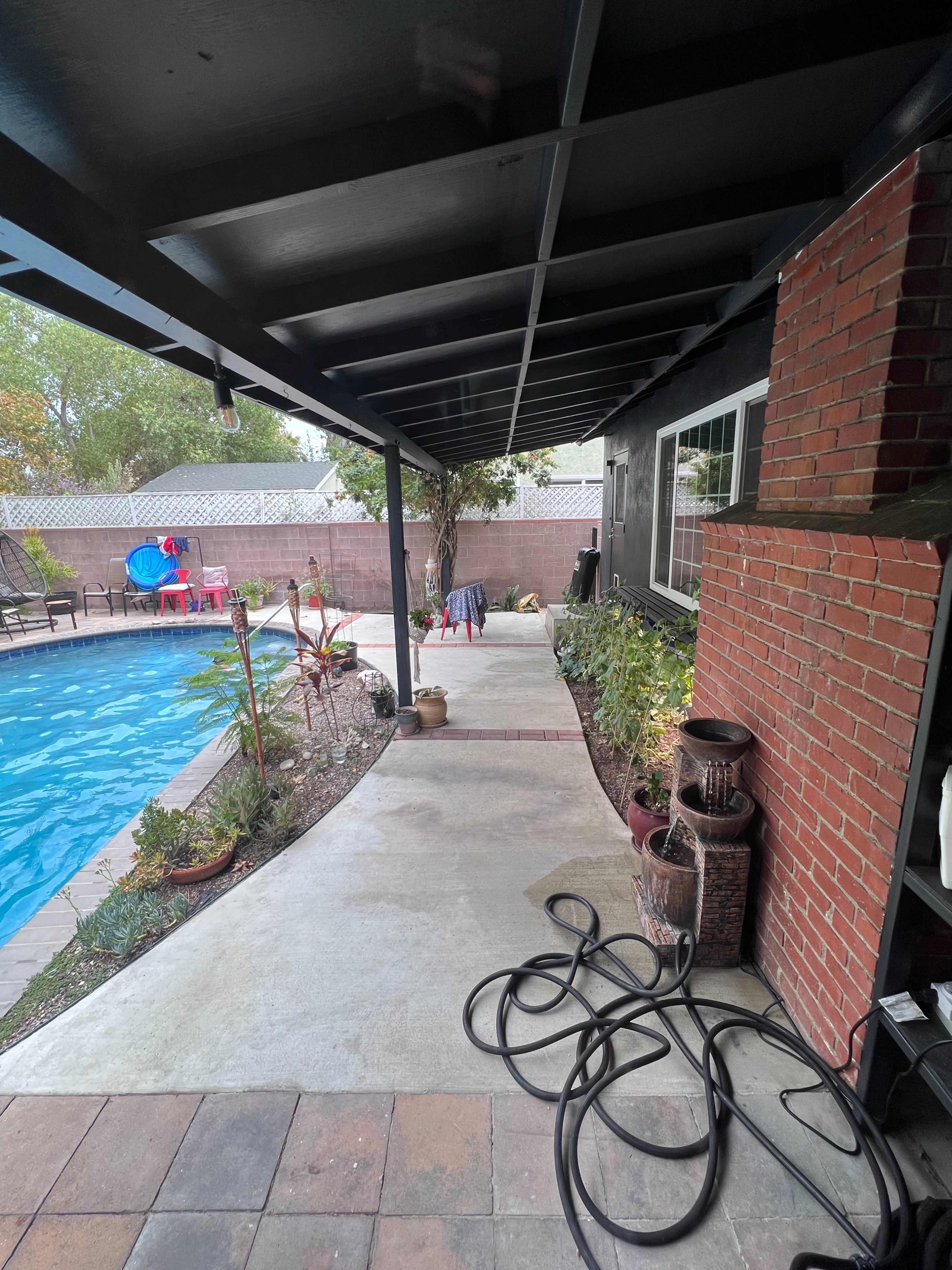 A concrete pathway runs alongside a swimming pool, flanked by plants and a brick wall under a covered patio.