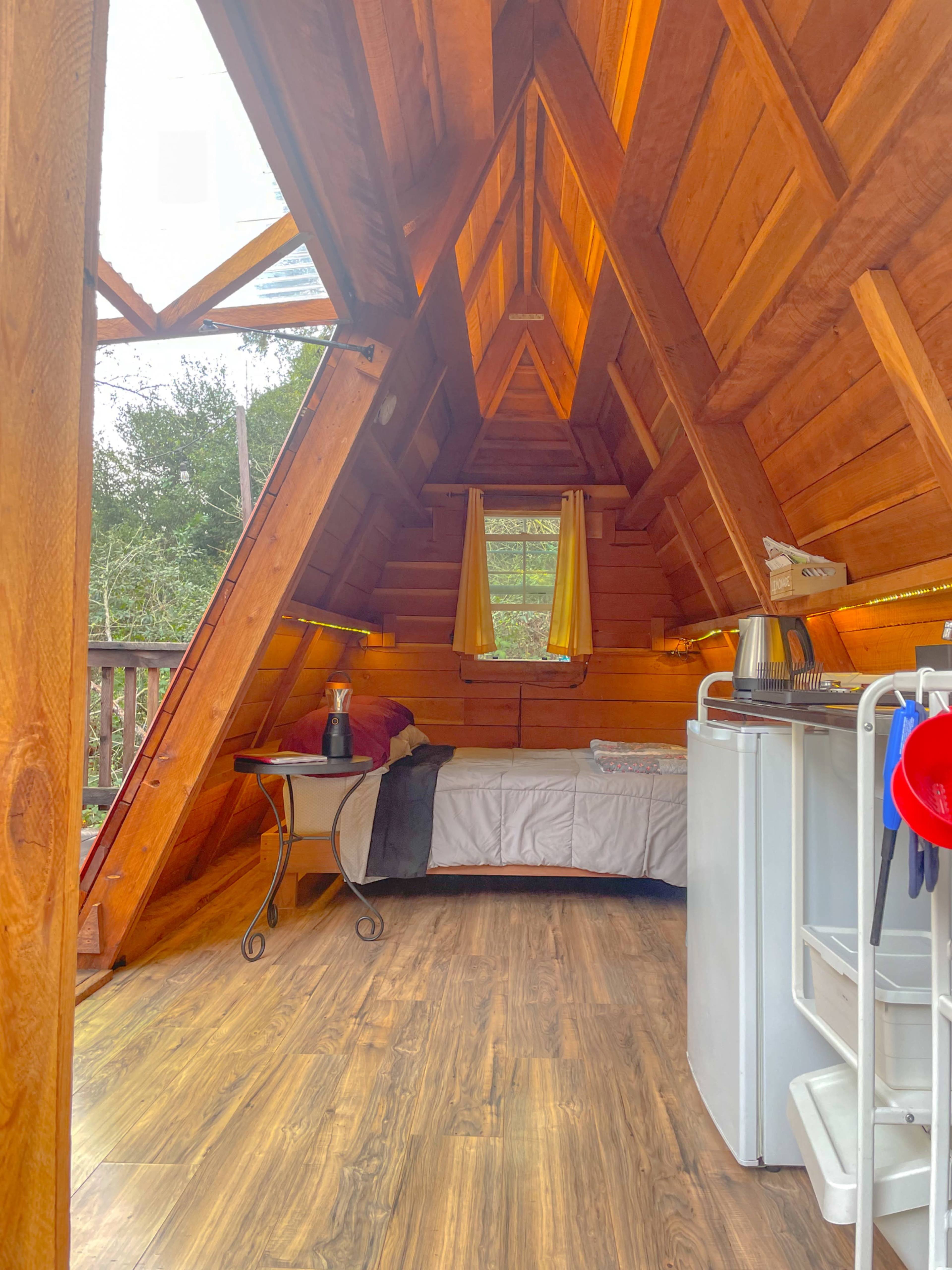 The interior of a wooden A-frame cabin features a bed, a small table, a kitchen area, and large windows with yellow curtains.