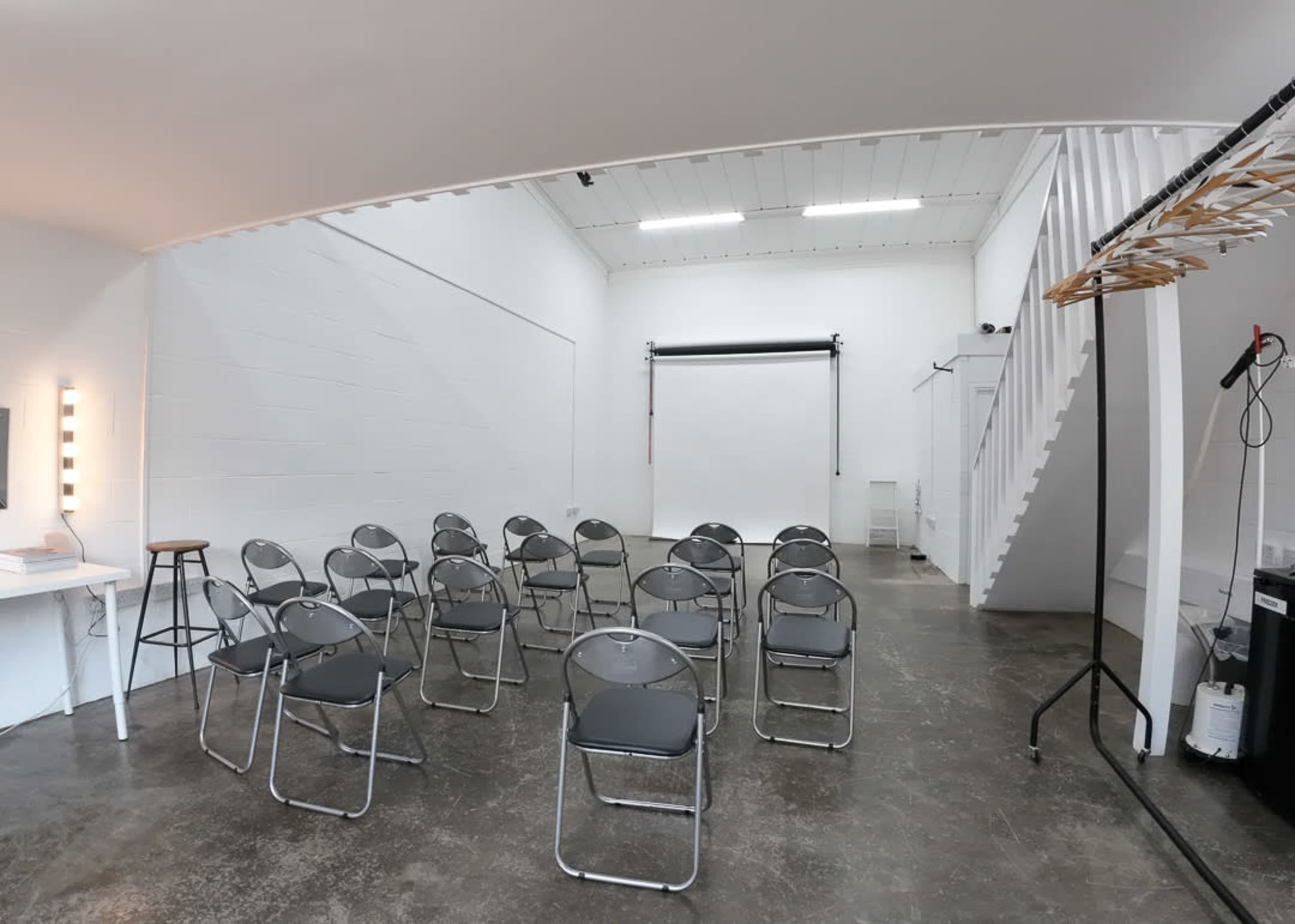 An empty room with metal folding chairs arranged in rows, a white wall, and a backdrop stand for photography.