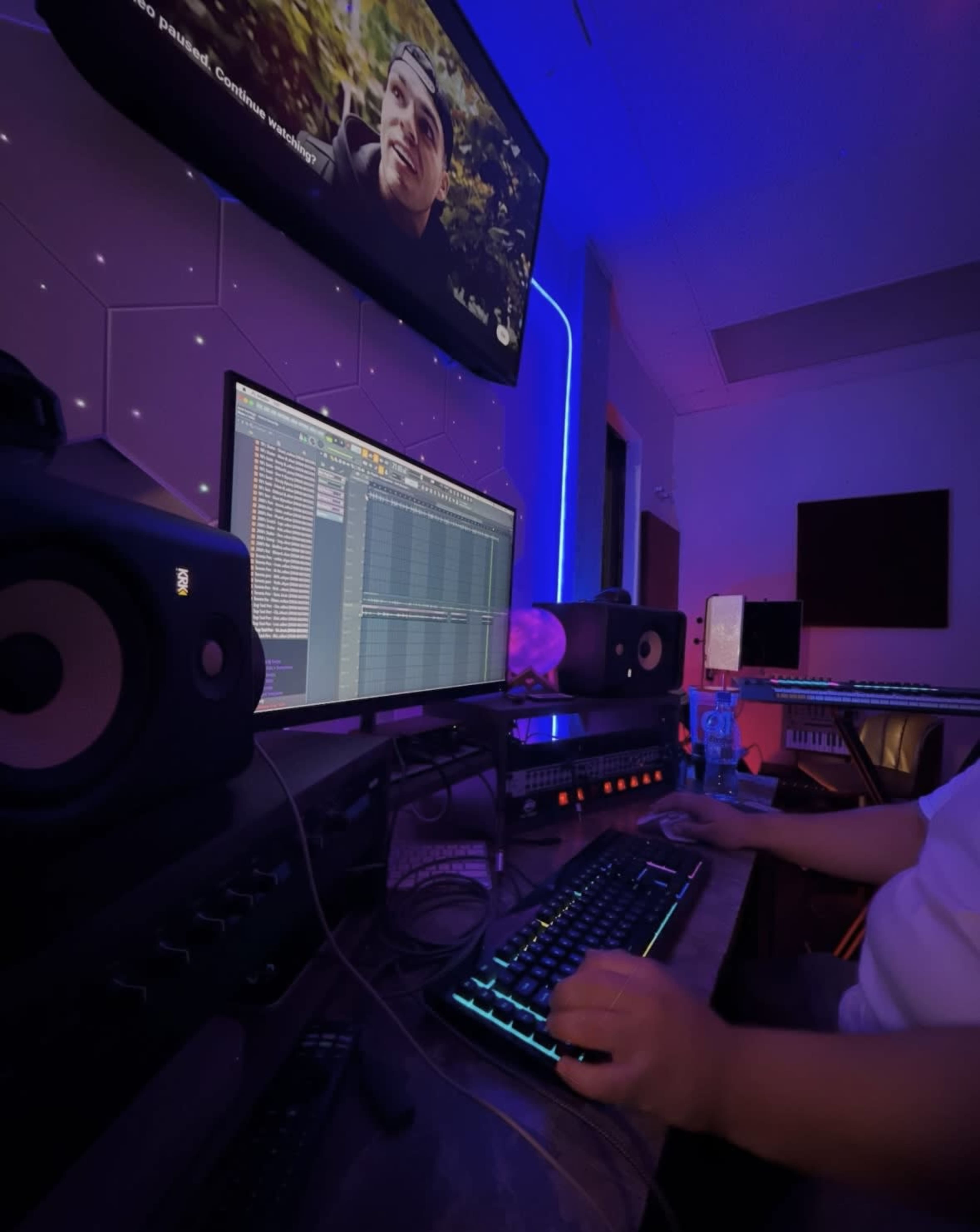 A person is working on a music production setup with multiple monitors, speakers, and ambient lighting in a dimly lit room.