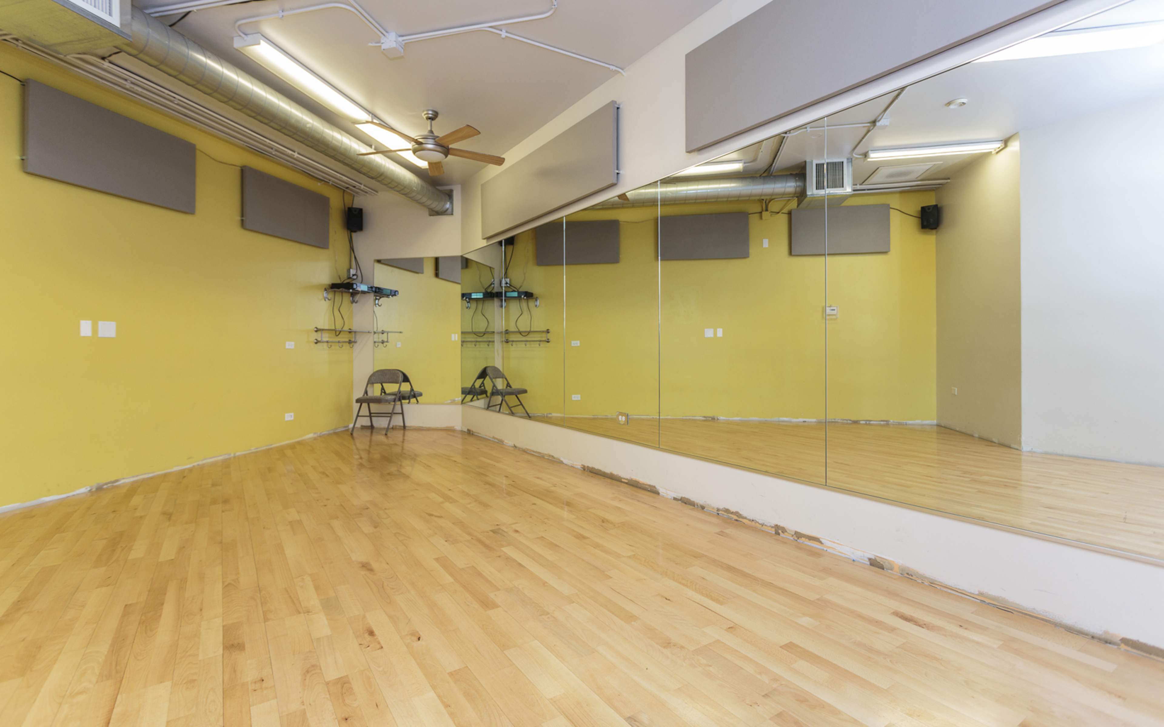 The image shows a spacious, empty dance studio featuring wooden flooring, large mirrors on one wall, and two folding chairs set against a yellow-painted wall.