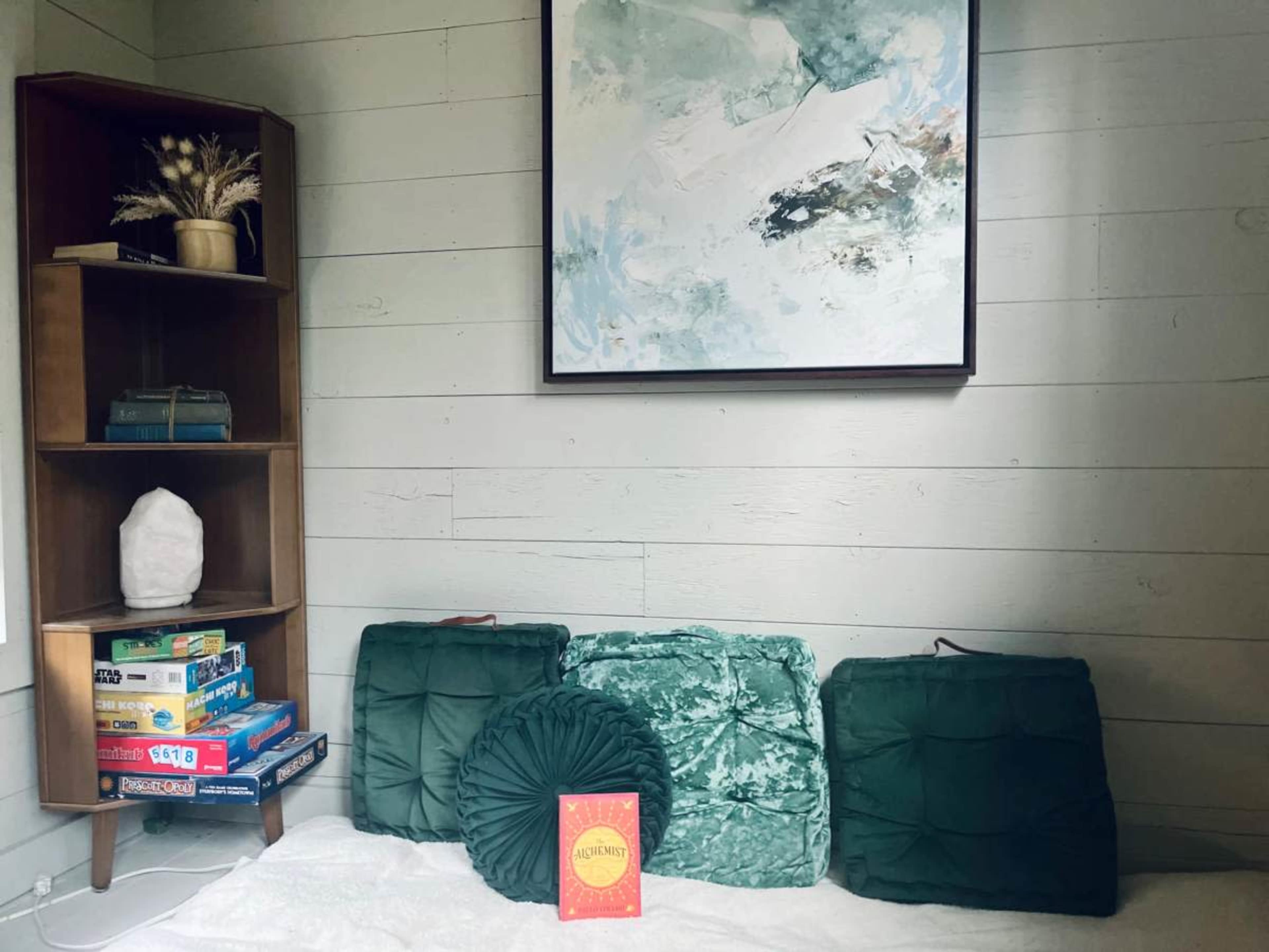 A cozy interior with a shelf stacked with board games, green cushions on a white surface, and an abstract painting on the wall.