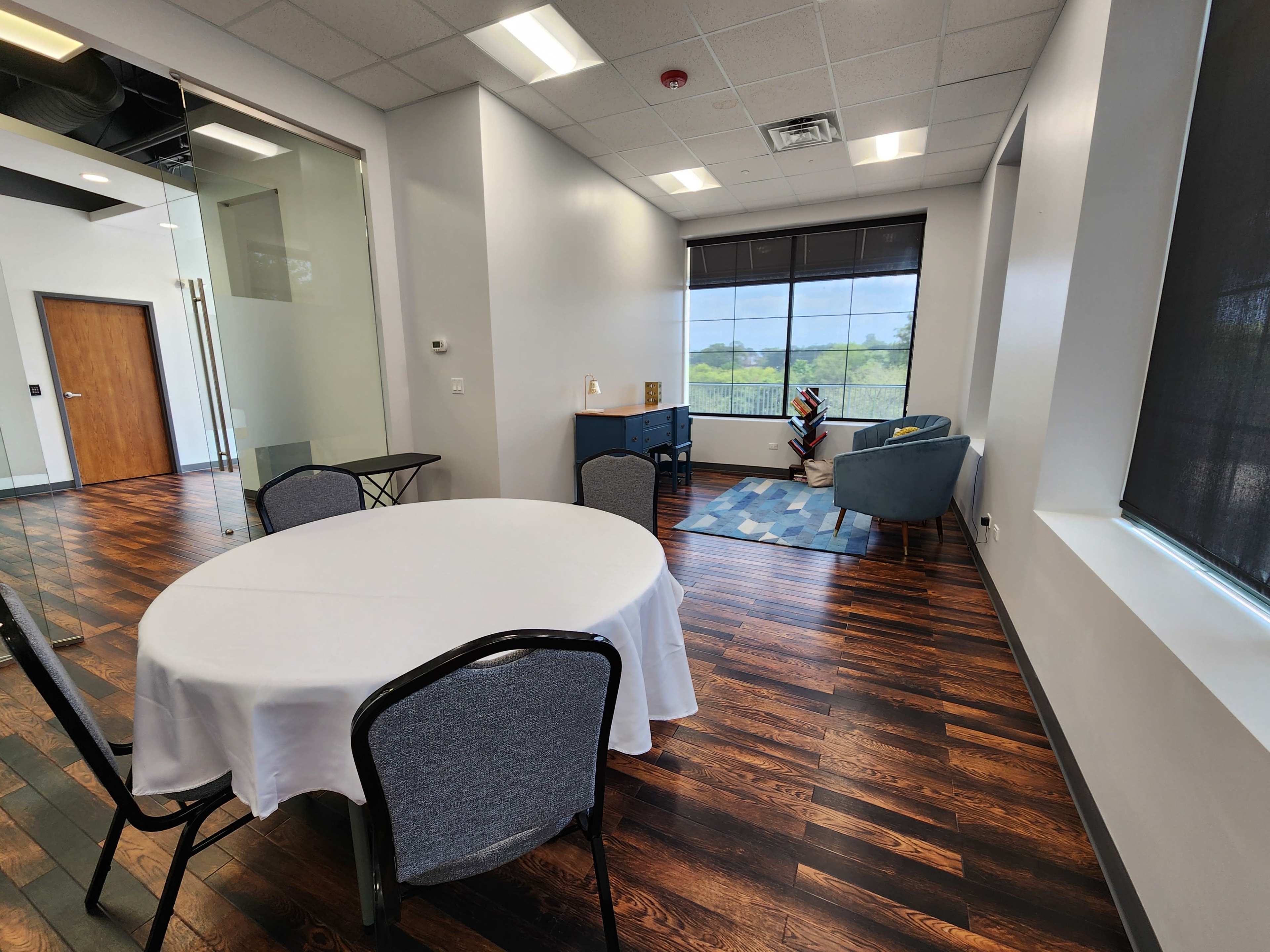 A bright, modern room with a round table covered by a white tablecloth, a blue armchair, and a large window overlooking greenery.