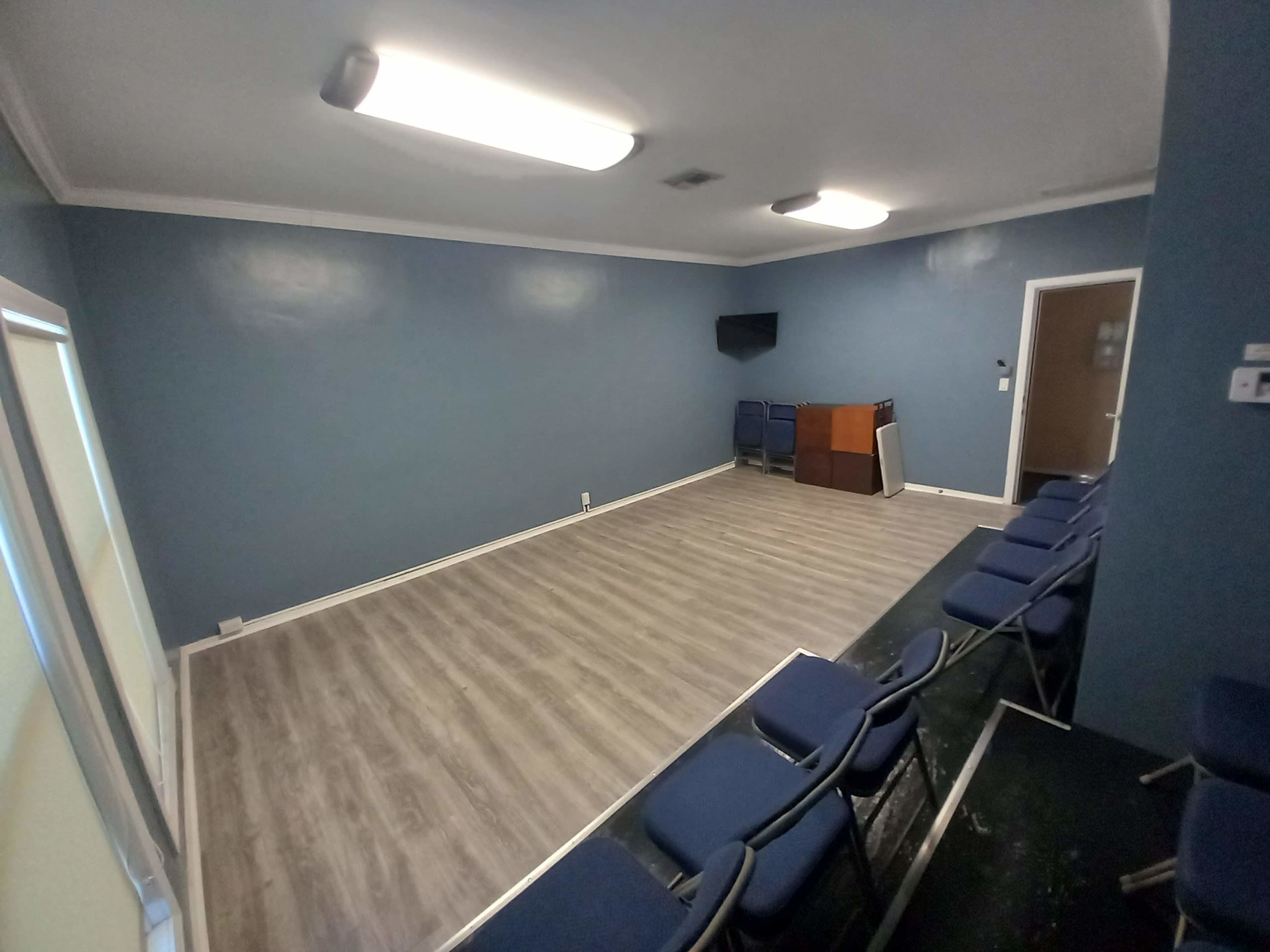 A simple, sparsely furnished room with a blue wall, a wooden podium, a television mounted on the wall, and several chairs arranged in rows.