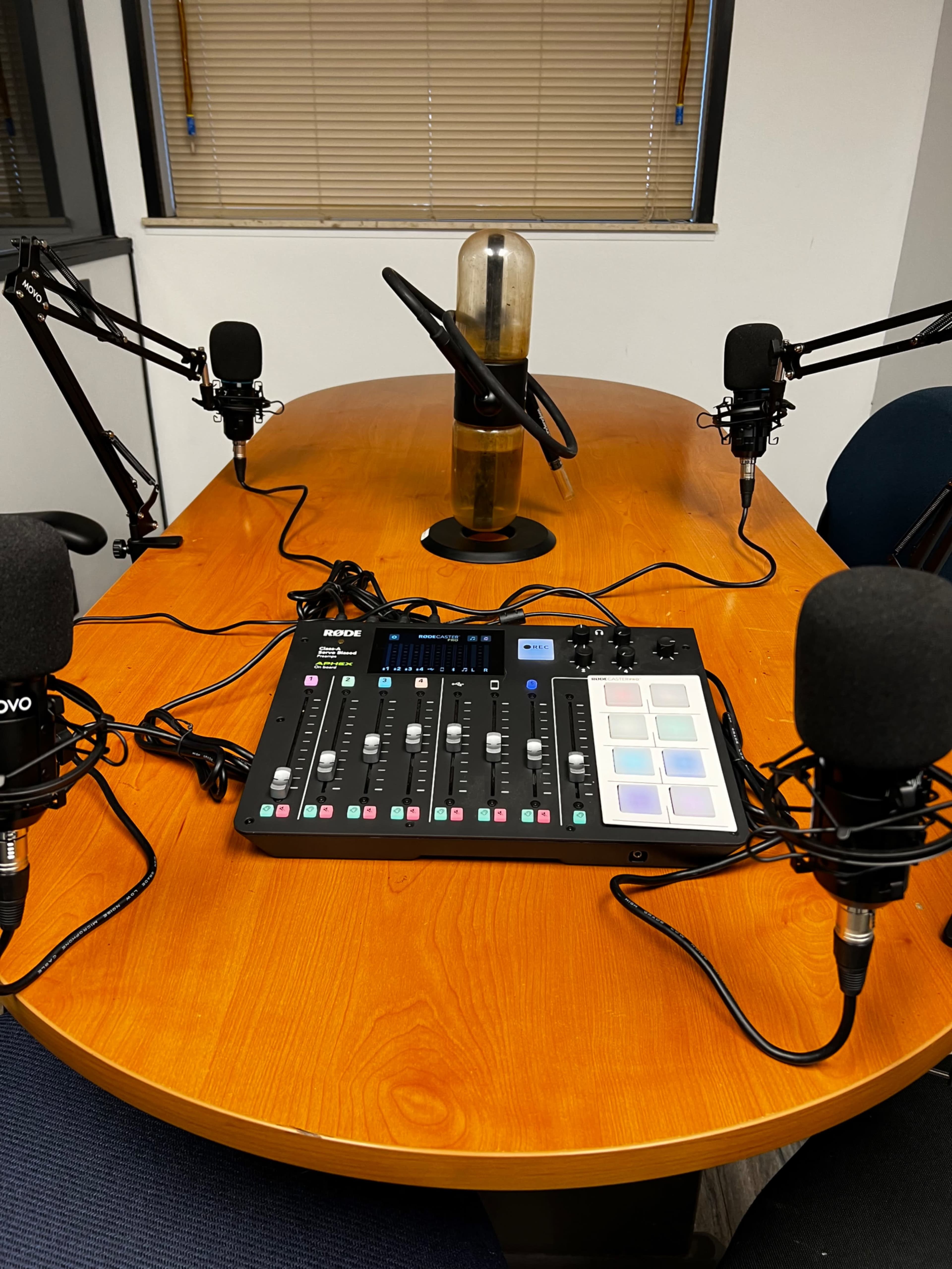 A wooden table is set up for a recording session, featuring a mixer, multiple microphones on adjustable arms, and a tall glass in the center.