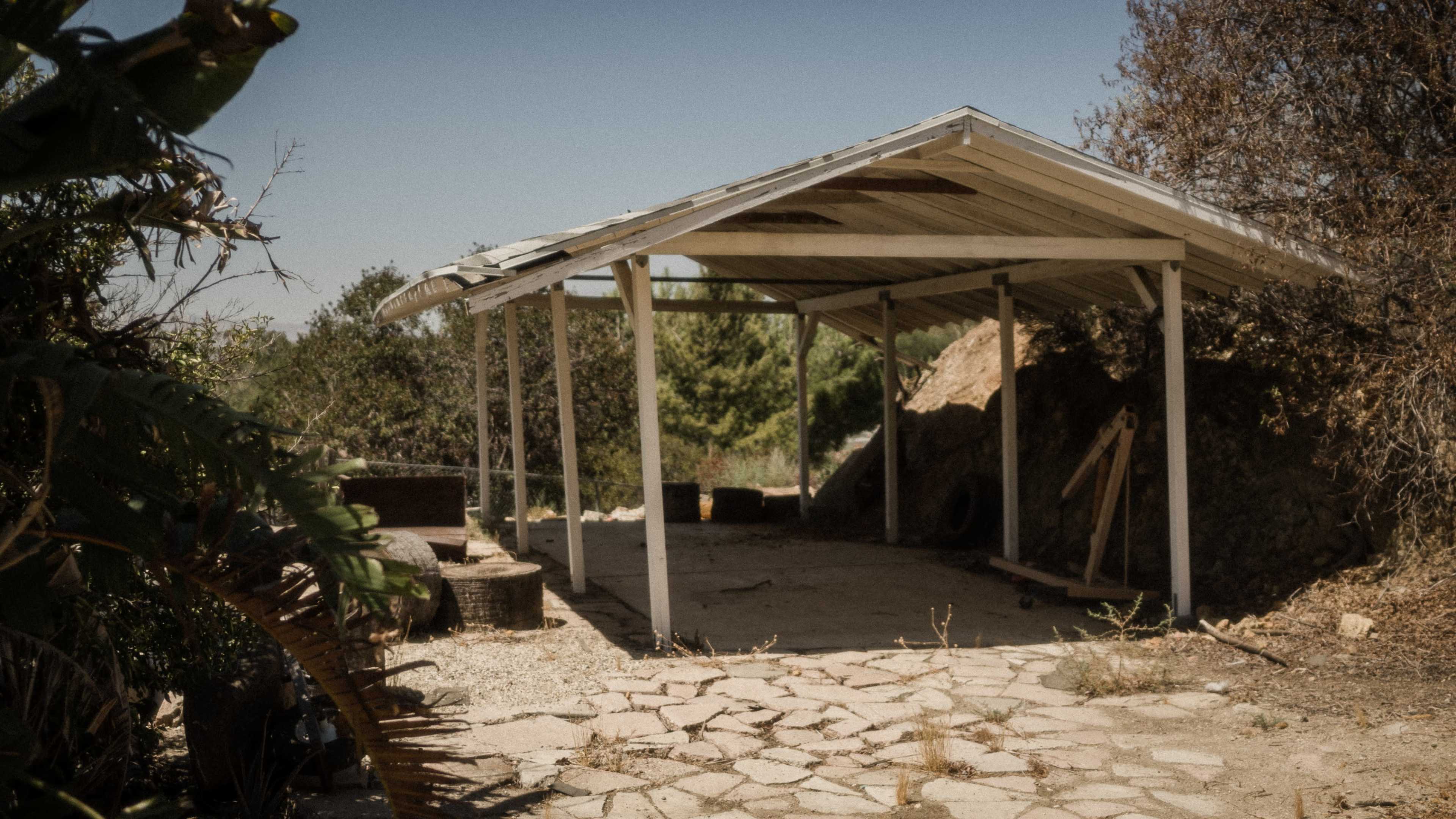 An open, roofed structure sits on a stone pathway surrounded by overgrown vegetation.