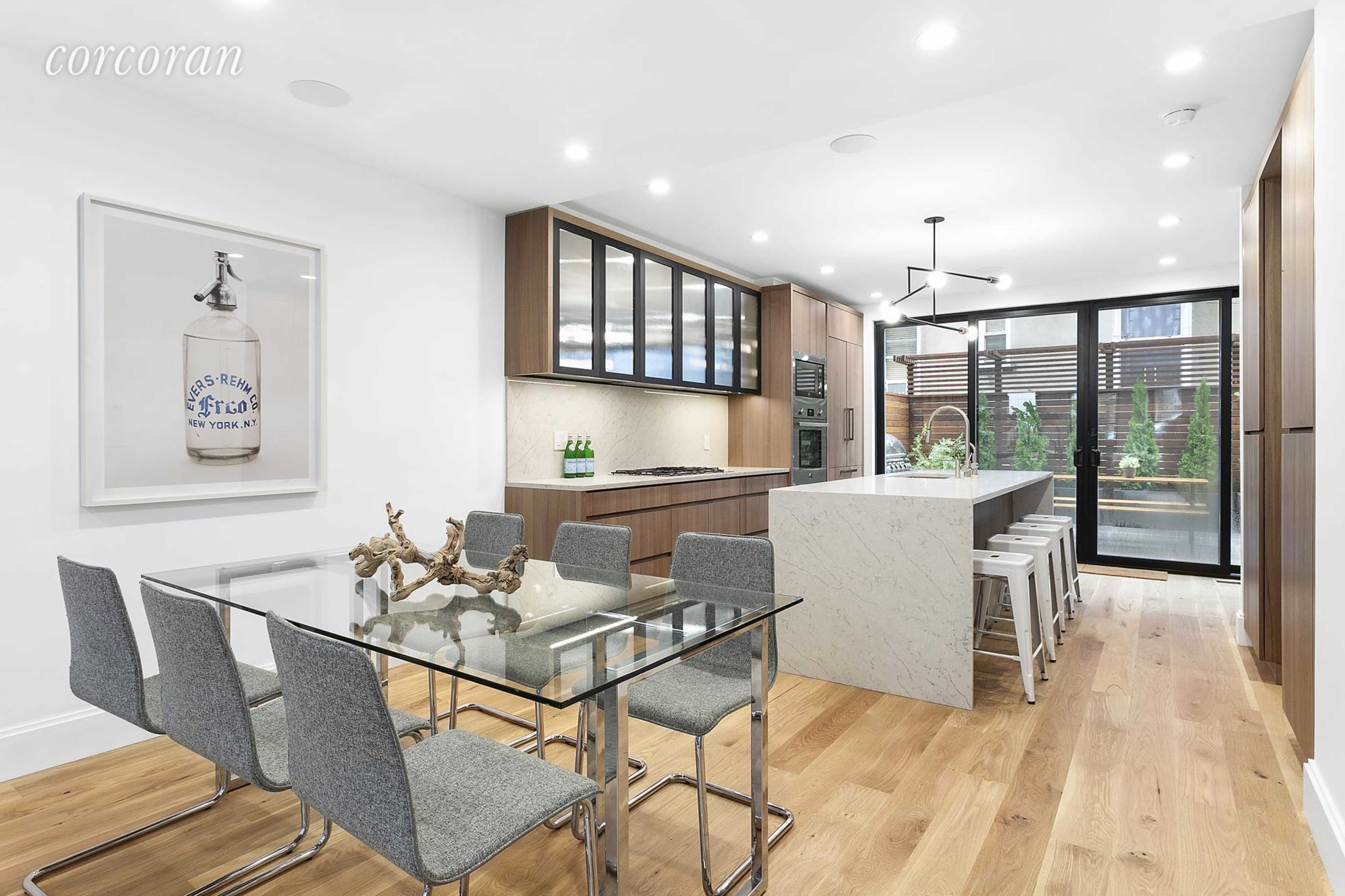 A modern kitchen and dining area featuring a glass table with six chairs, an island with a marble countertop, and large windows providing natural light.
