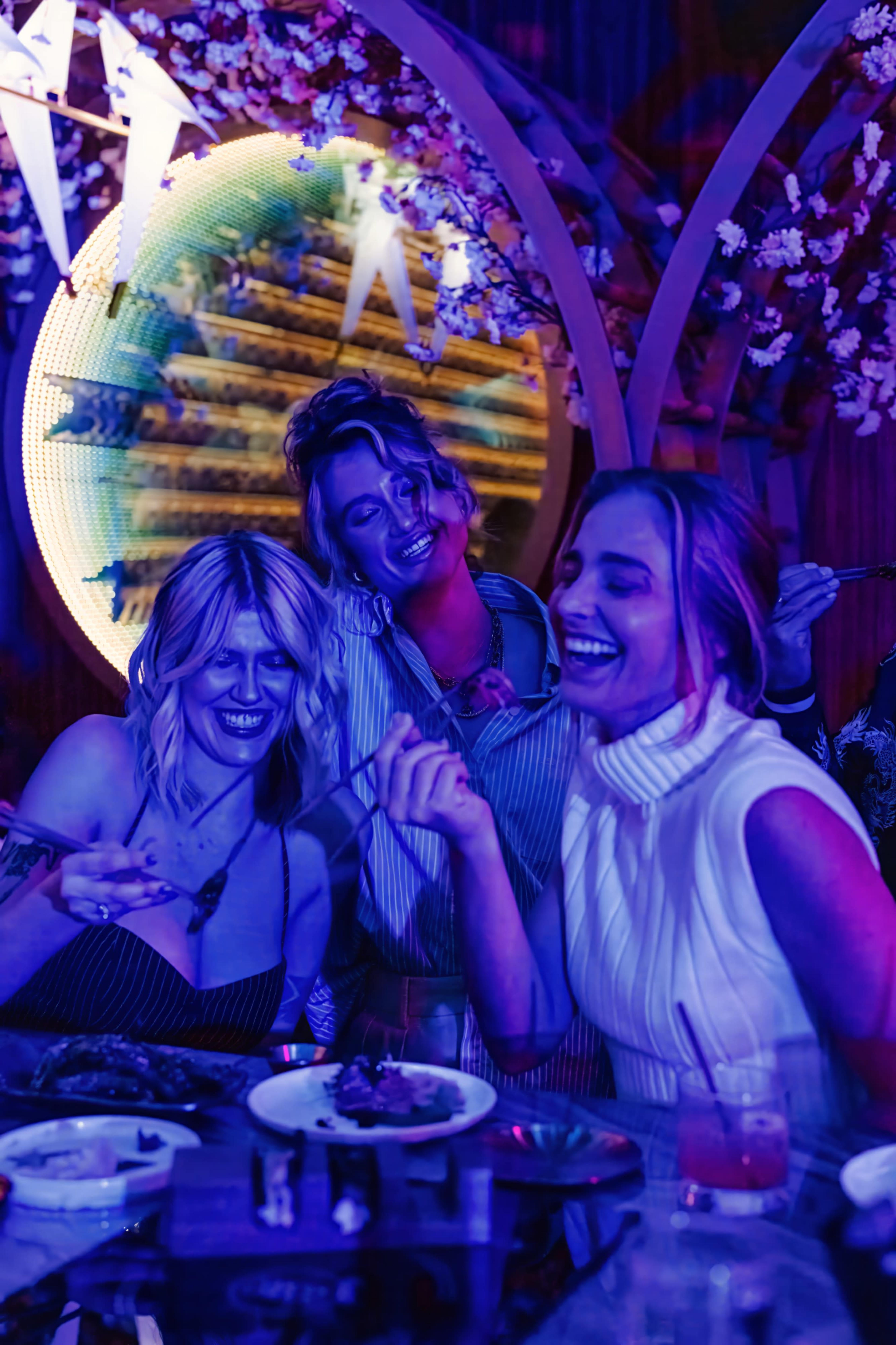 Three women are enjoying a lively dinner party, laughing and sharing food under colorful lighting and decorative elements.