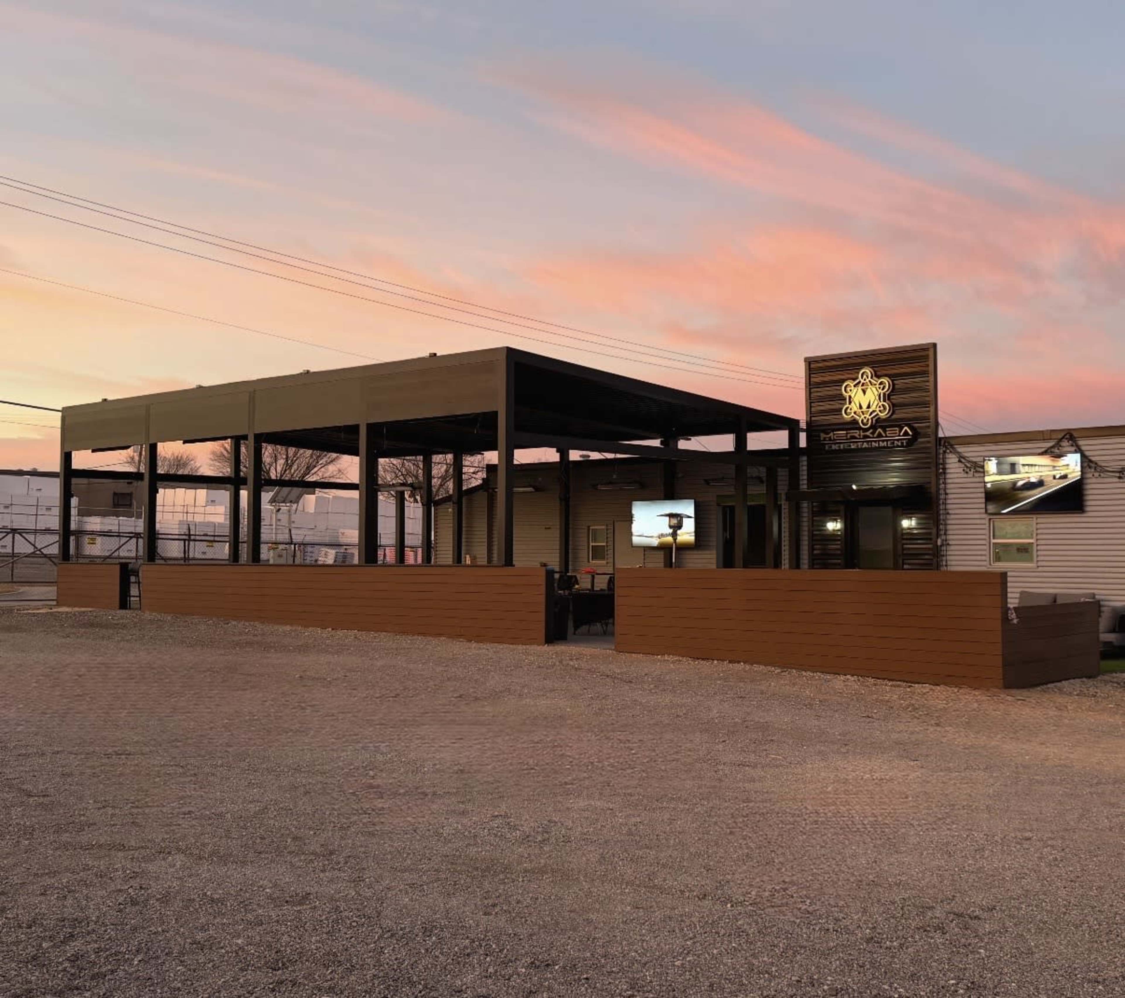 A modern building with a large outdoor seating area and screens, set against a colorful sunset sky.