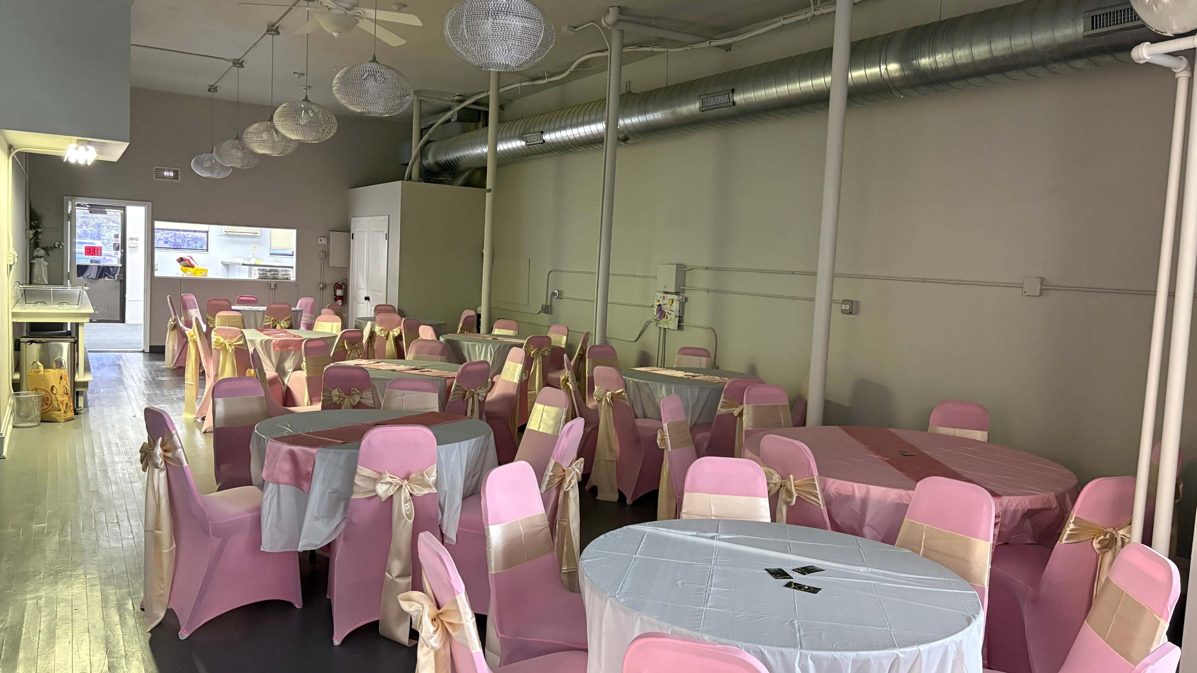 The image shows a banquet hall with several round tables covered in white and pastel pink tablecloths, surrounded by chairs adorned with pink and gold sashes.