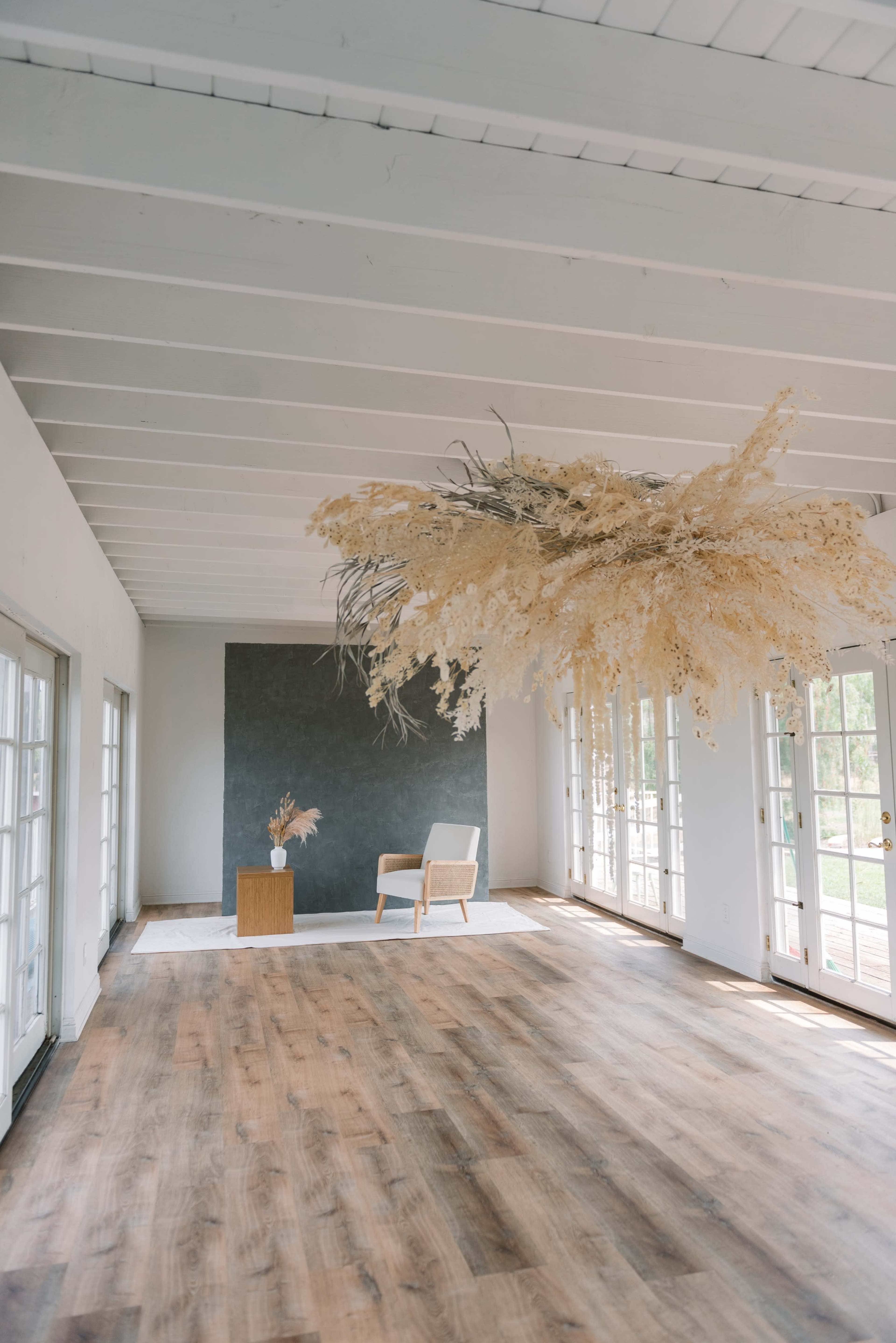 A minimalist interior room with wooden floors, large windows, and a striking dried floral arrangement suspended from the ceiling above a simple wooden side table and an armchair.