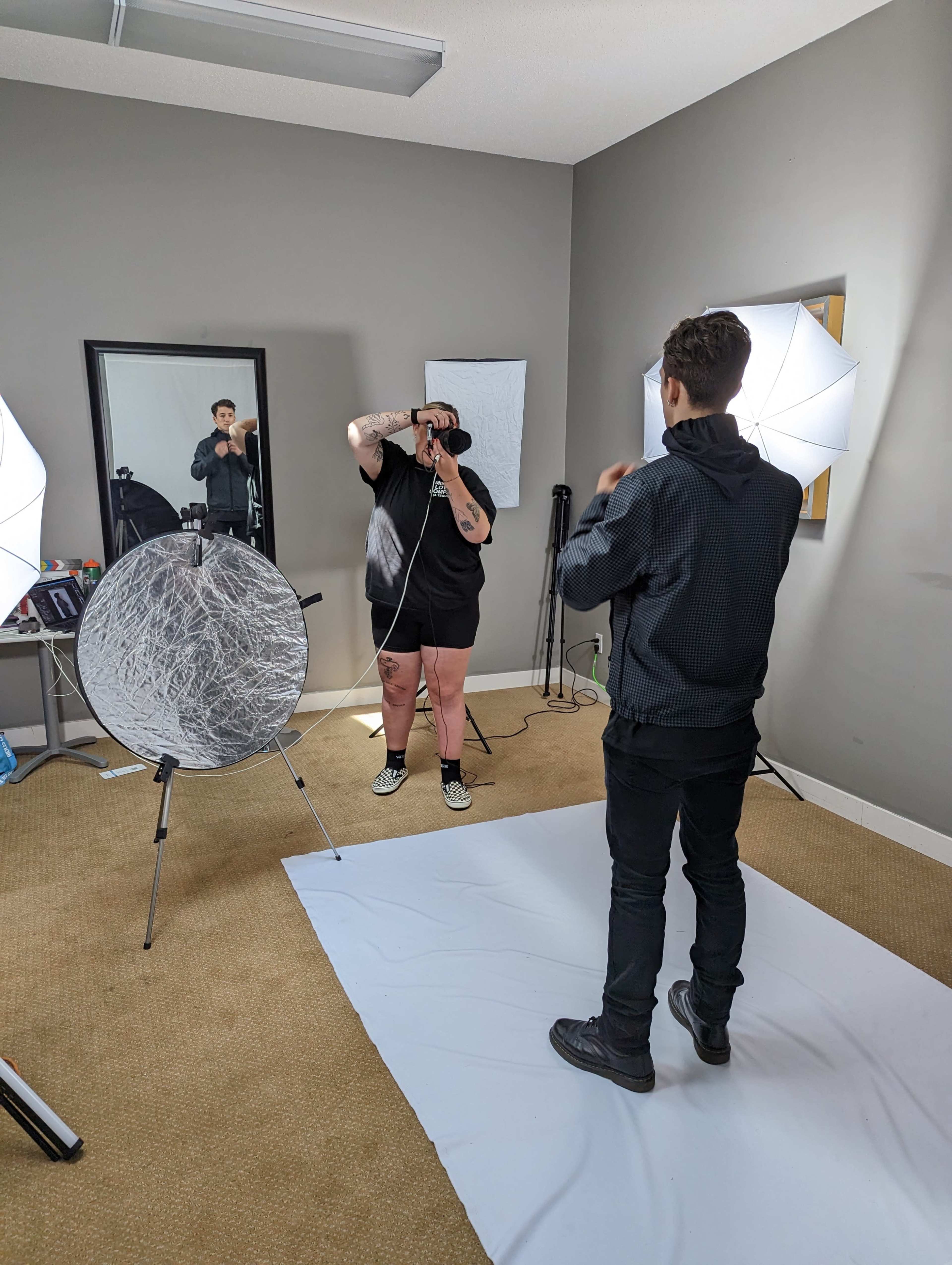 A person with tattoos stands in front of a mirror while another person adjusts equipment in a photo studio setup.