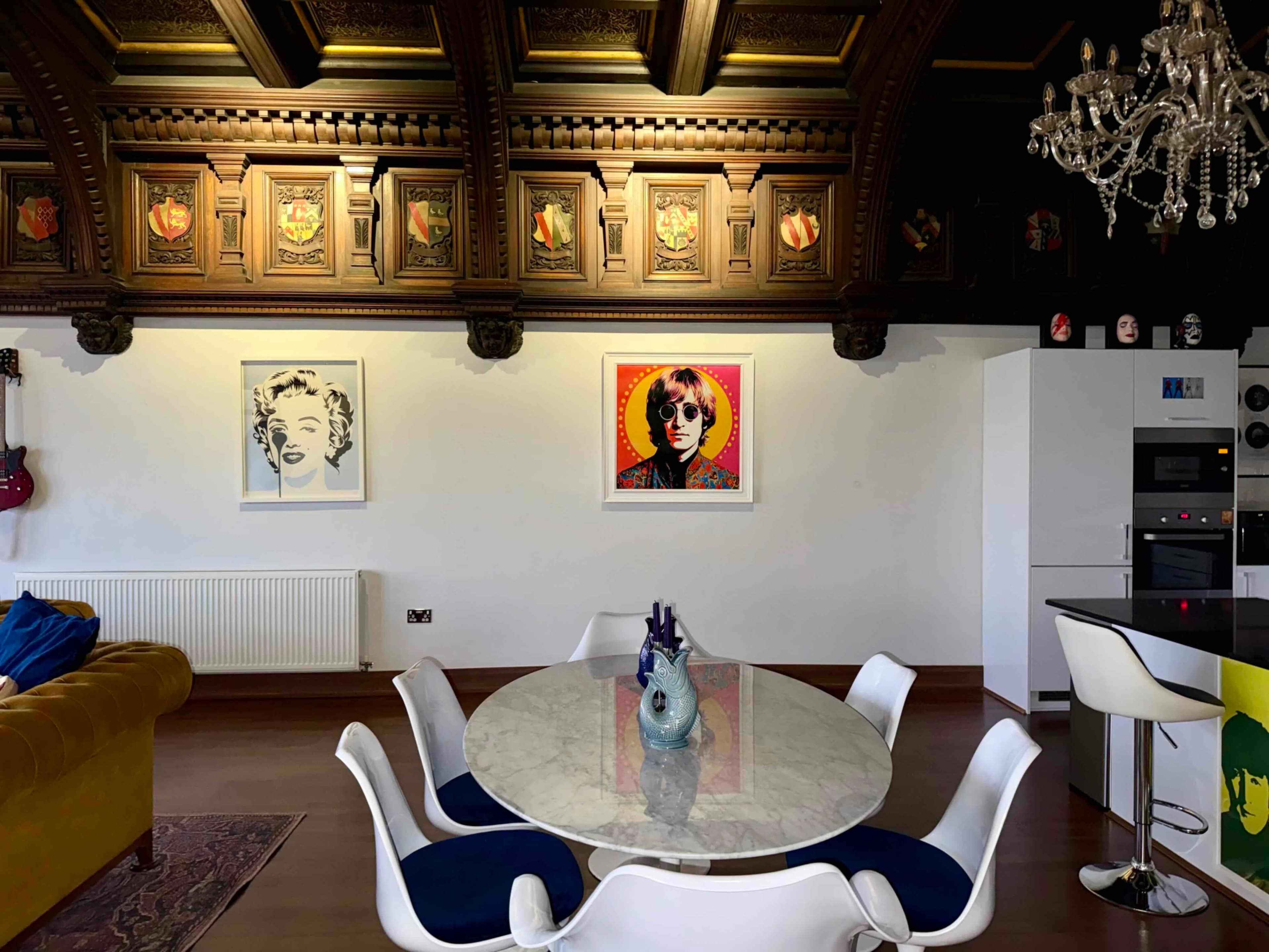 The image shows a modern dining area featuring a round marble table surrounded by white chairs, with vibrant artwork on the walls and ornate wooden panels above.