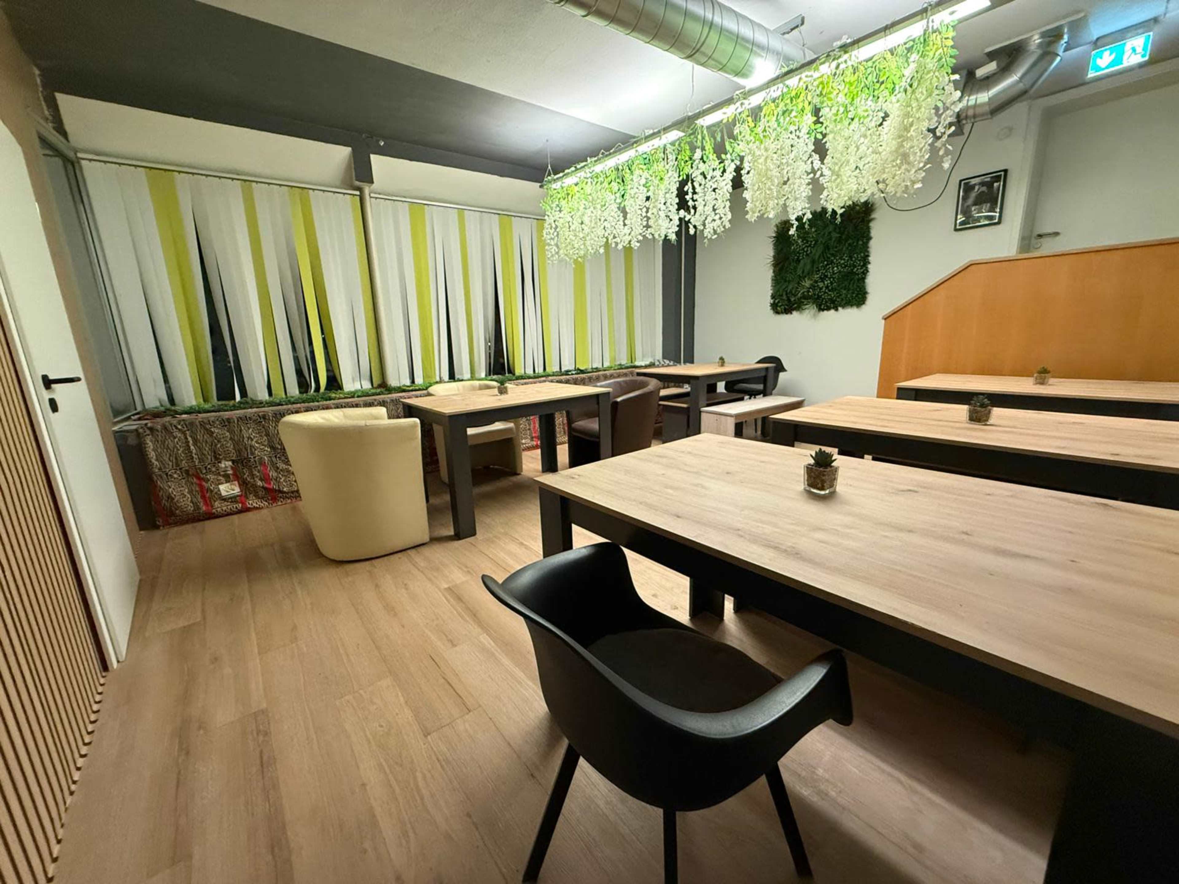 The image shows a modern indoor dining area with wooden tables, a mix of seating options, and decorative plants hanging from the ceiling.