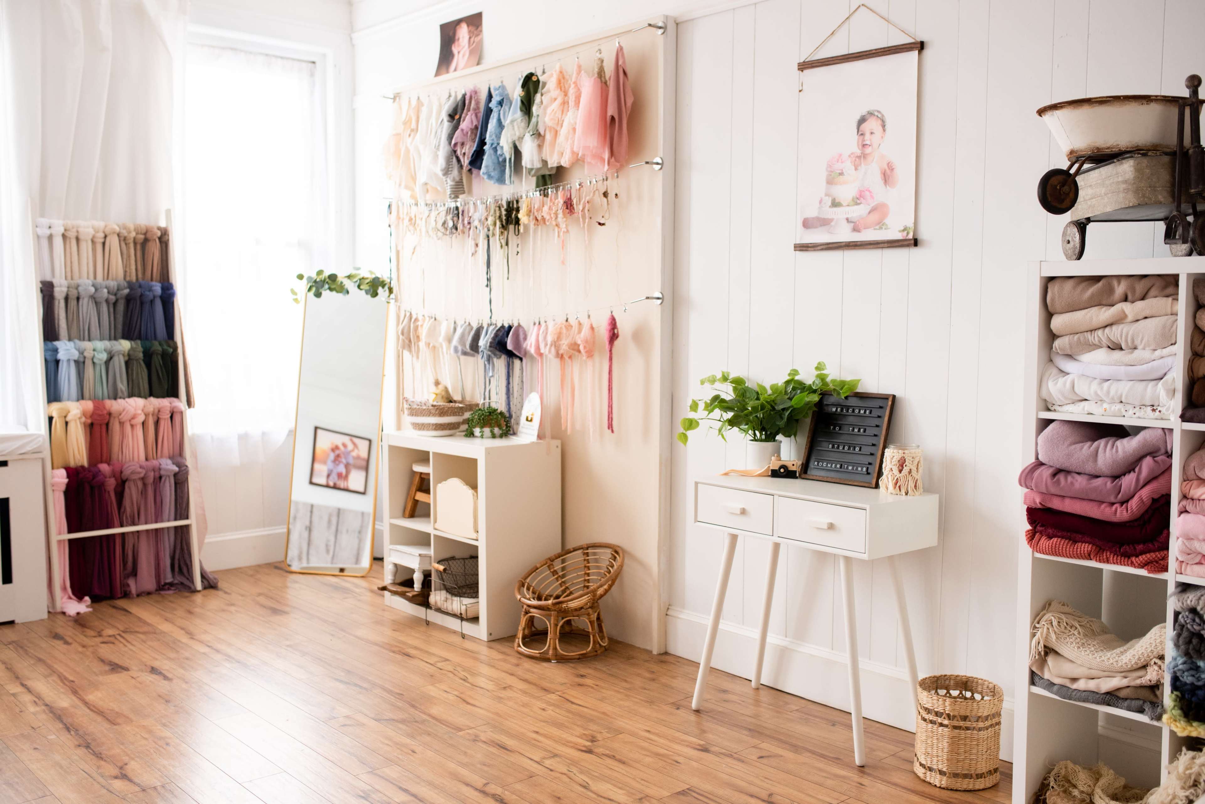 A bright, organized studio space with shelves displaying various fabric bundles, a large mirror, and decorative items, including a framed photo and a plant on a white table.