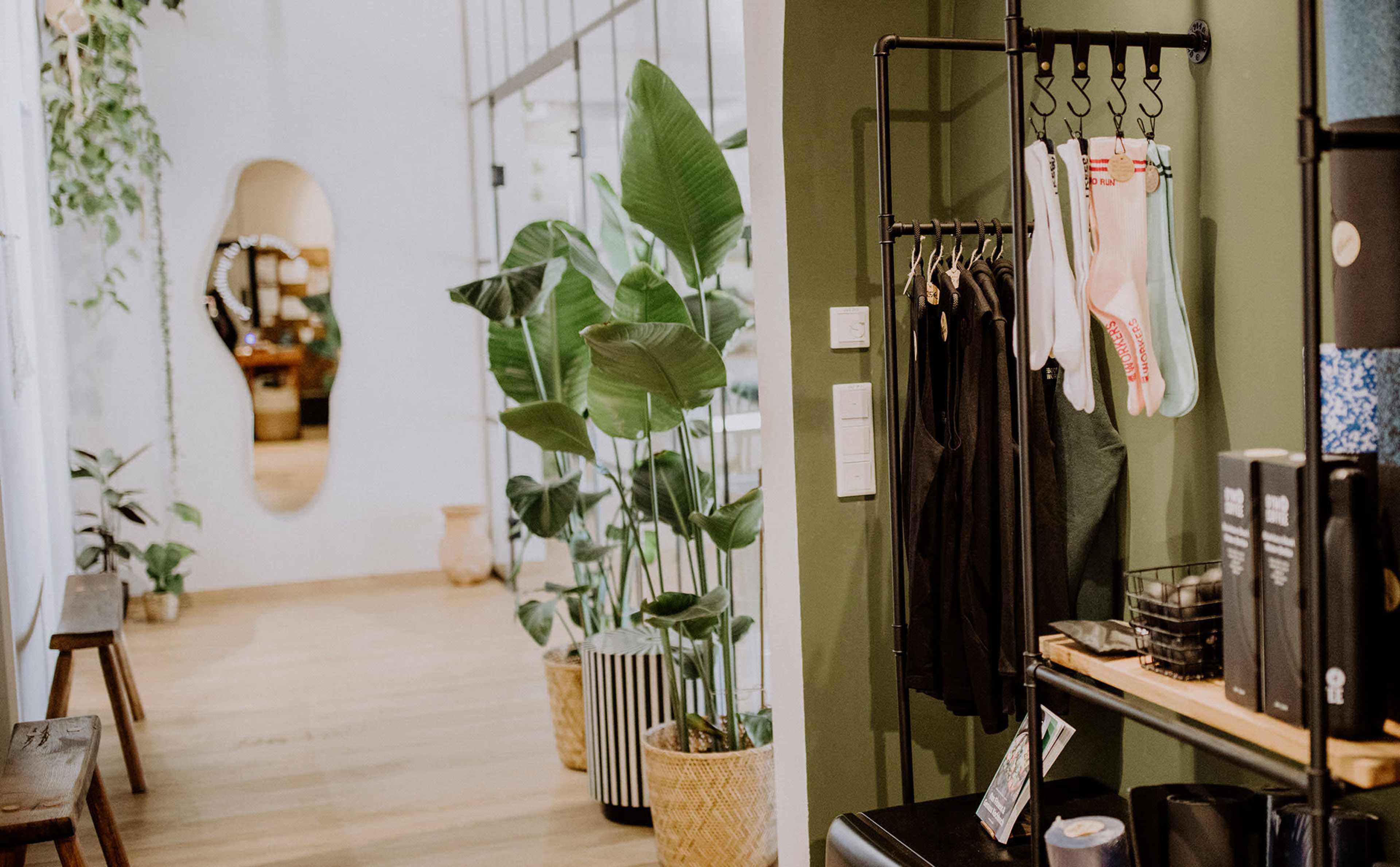 The image shows a stylish interior space with green plants, a mirrored wall, and clothing and accessories displayed on racks.