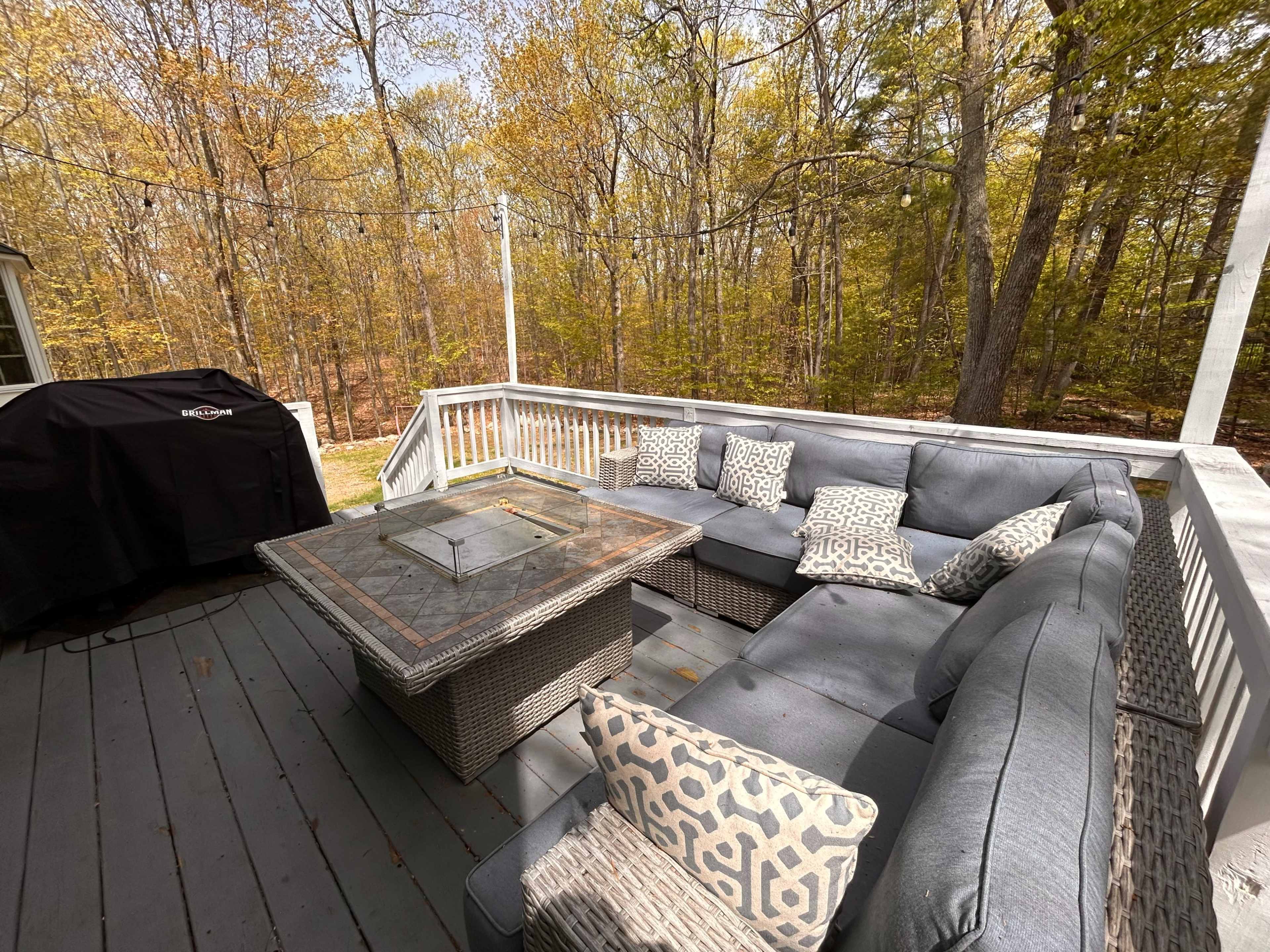A spacious outdoor patio features a sectional sofa with decorative pillows and a square fire pit, surrounded by trees with autumn foliage.