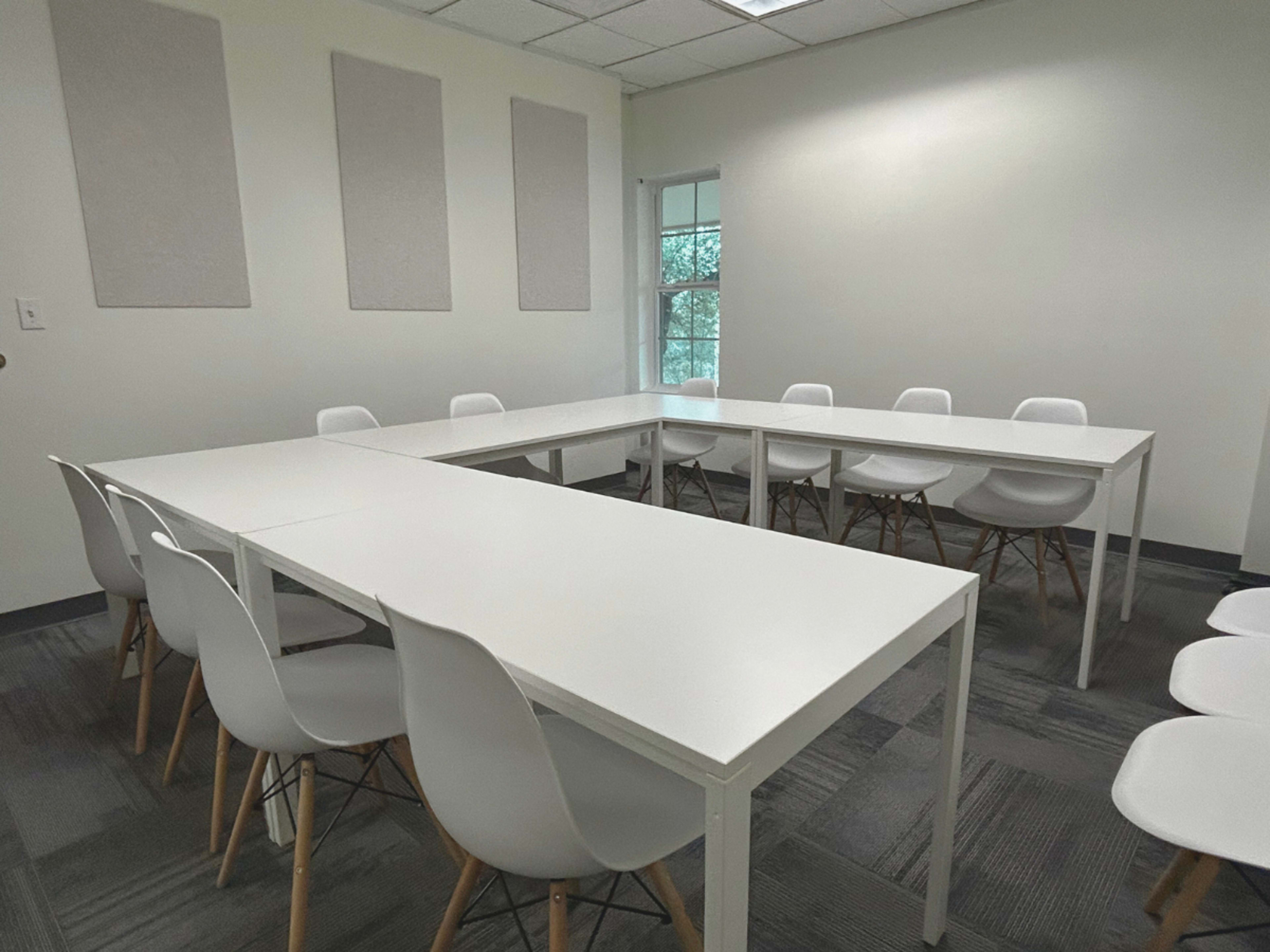 A conference room is set up with several white tables arranged in a U-shape and surrounded by white chairs.