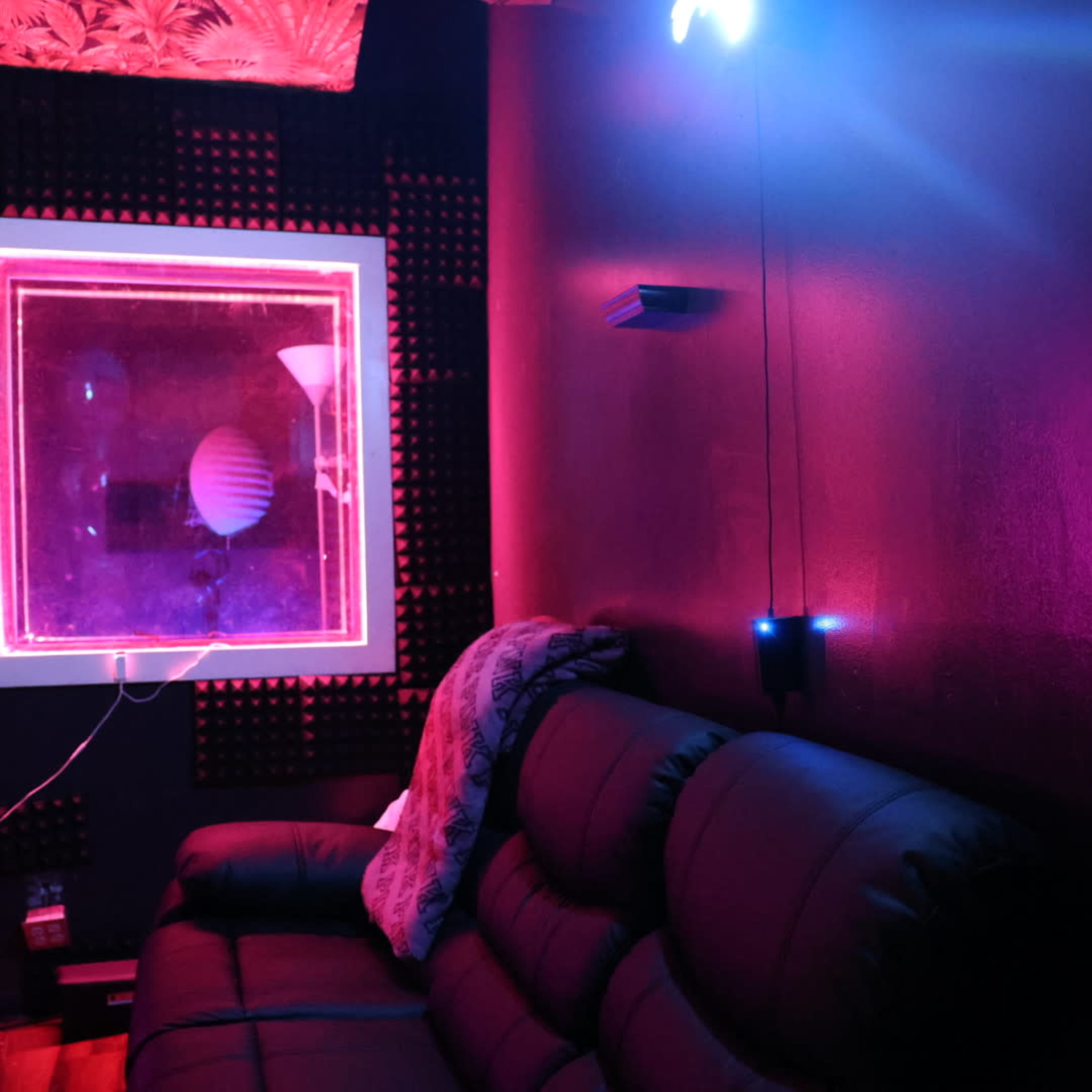 The image shows a dark room with a black sofa, illuminated by purple and pink lights, and featuring a window with a neon frame.