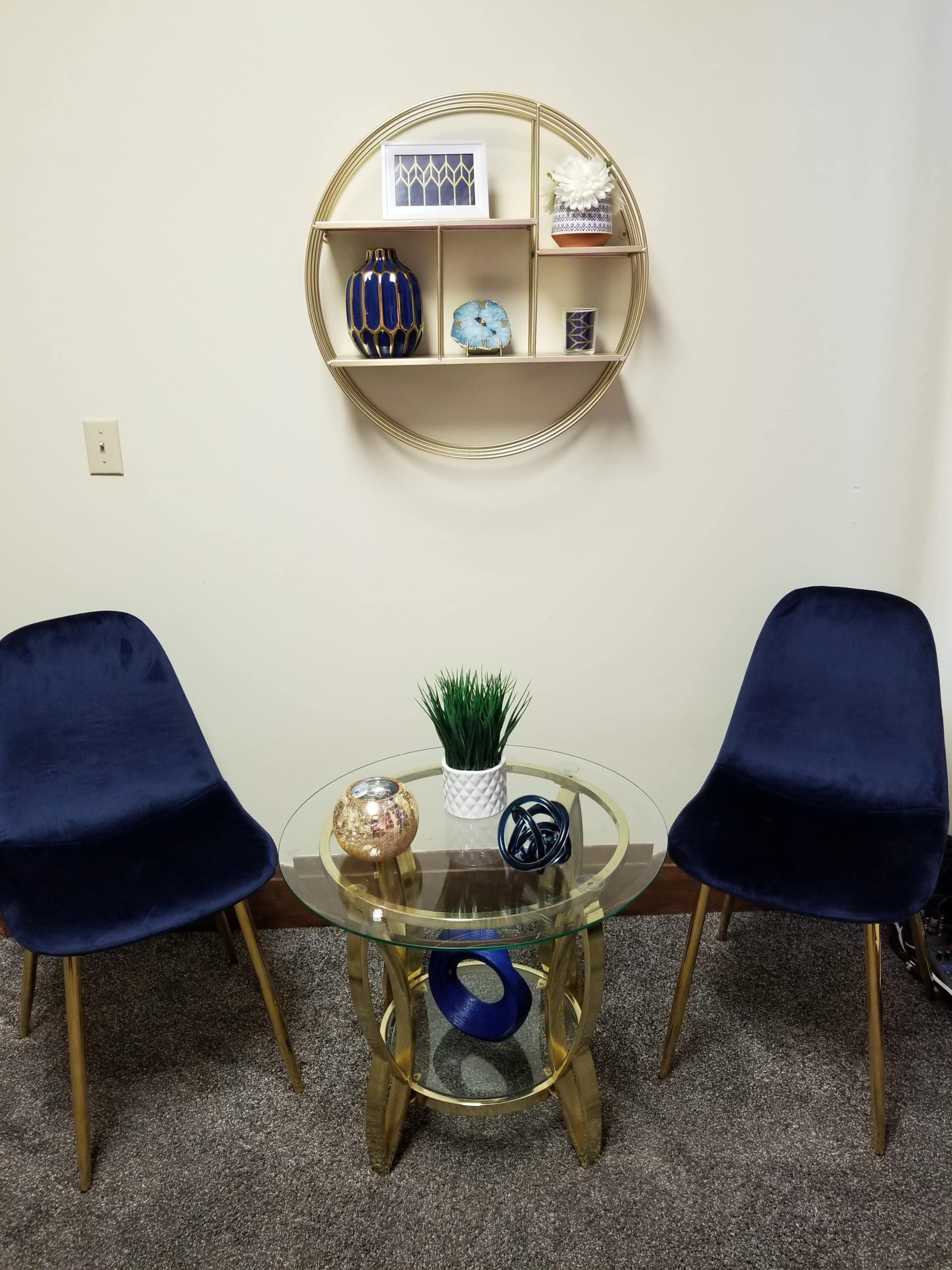 A small sitting area features two dark blue chairs flanking a glass table, with a decorative shelf mounted on the wall above.