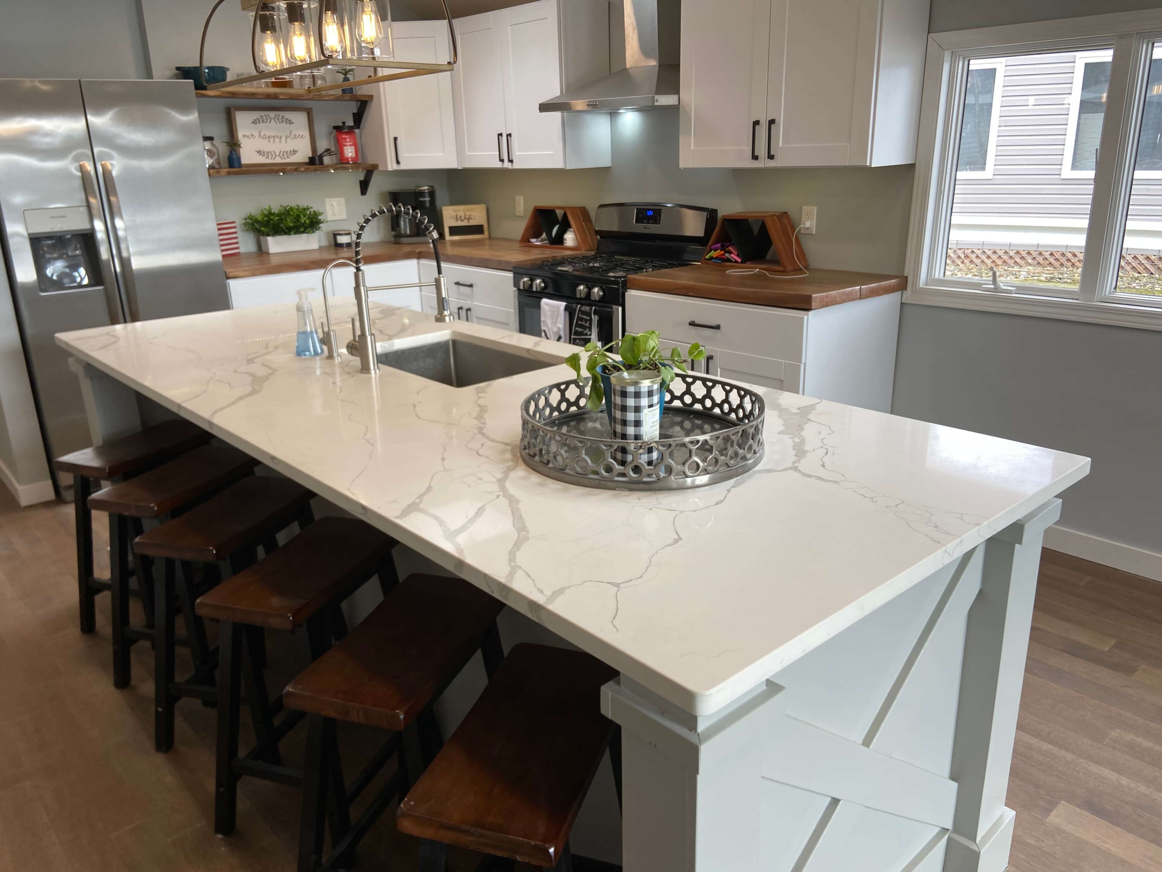 A modern kitchen features a large white countertop with a sink, surrounded by dark wooden stools, and equipped with stainless steel appliances.