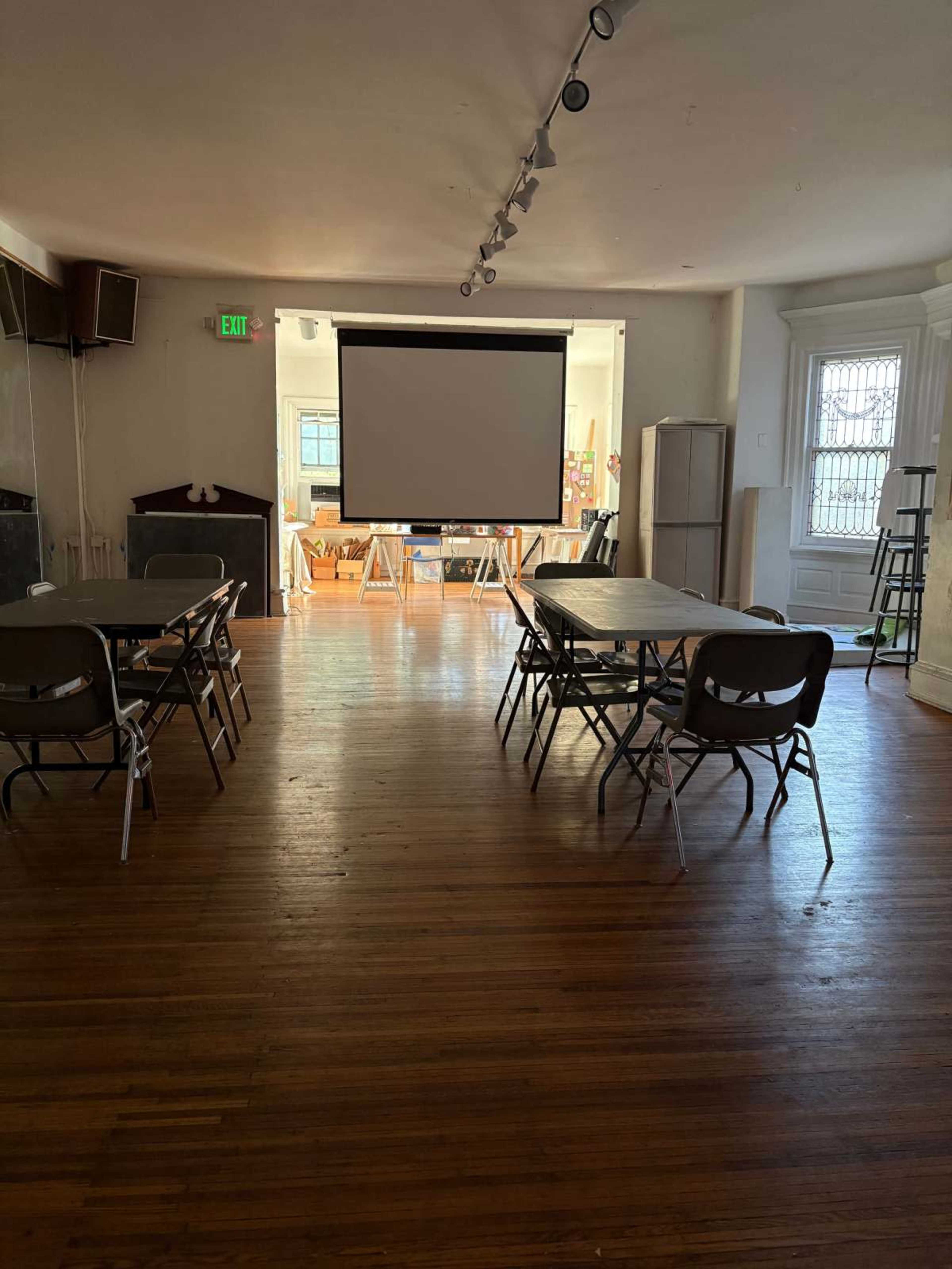 The image shows a spacious room with wooden floors, several folding tables and chairs arranged, and a projector screen at the front.