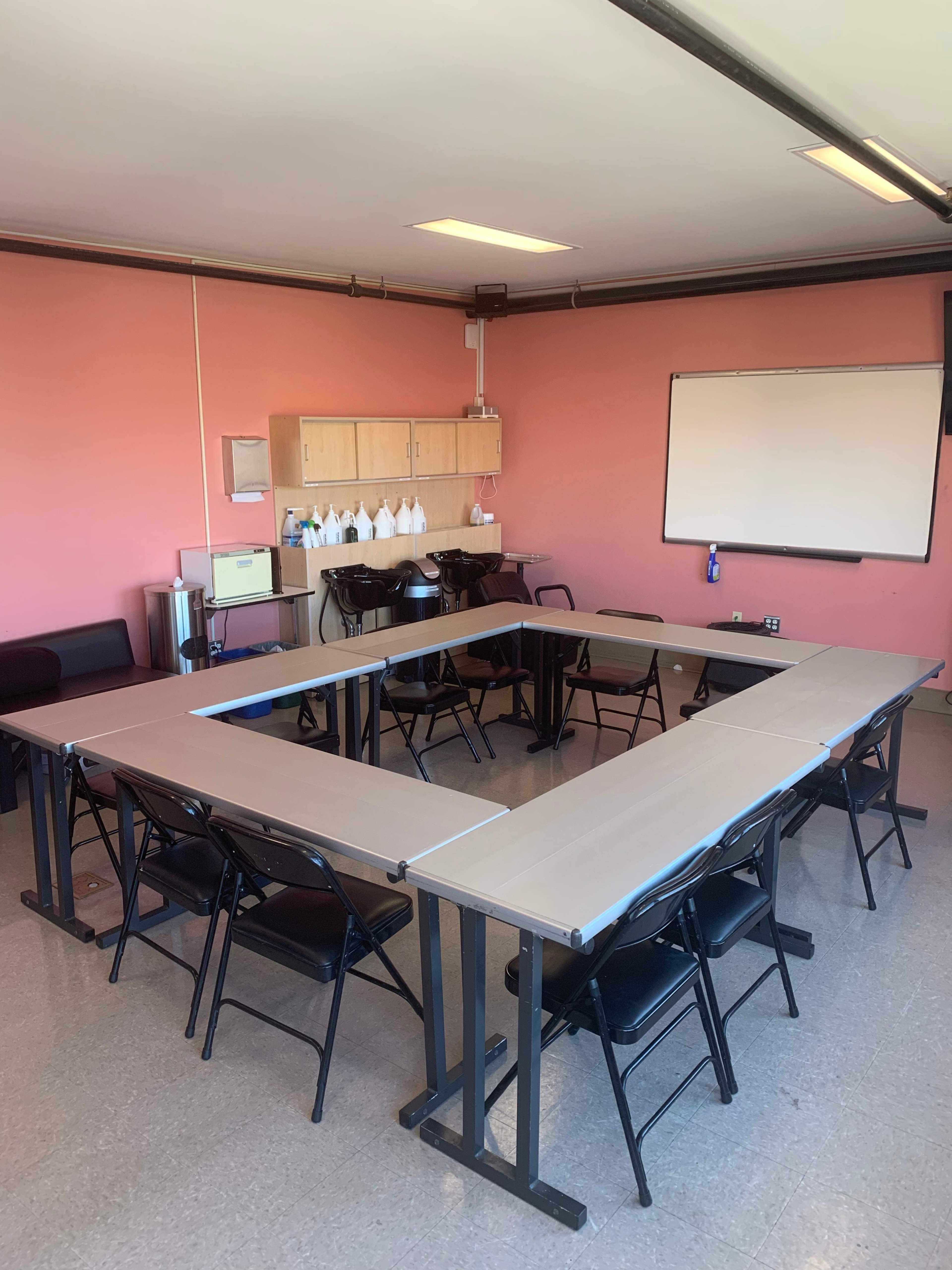 A meeting room features a rectangular arrangement of tables surrounded by black chairs, with a pink wall and a whiteboard in the background.