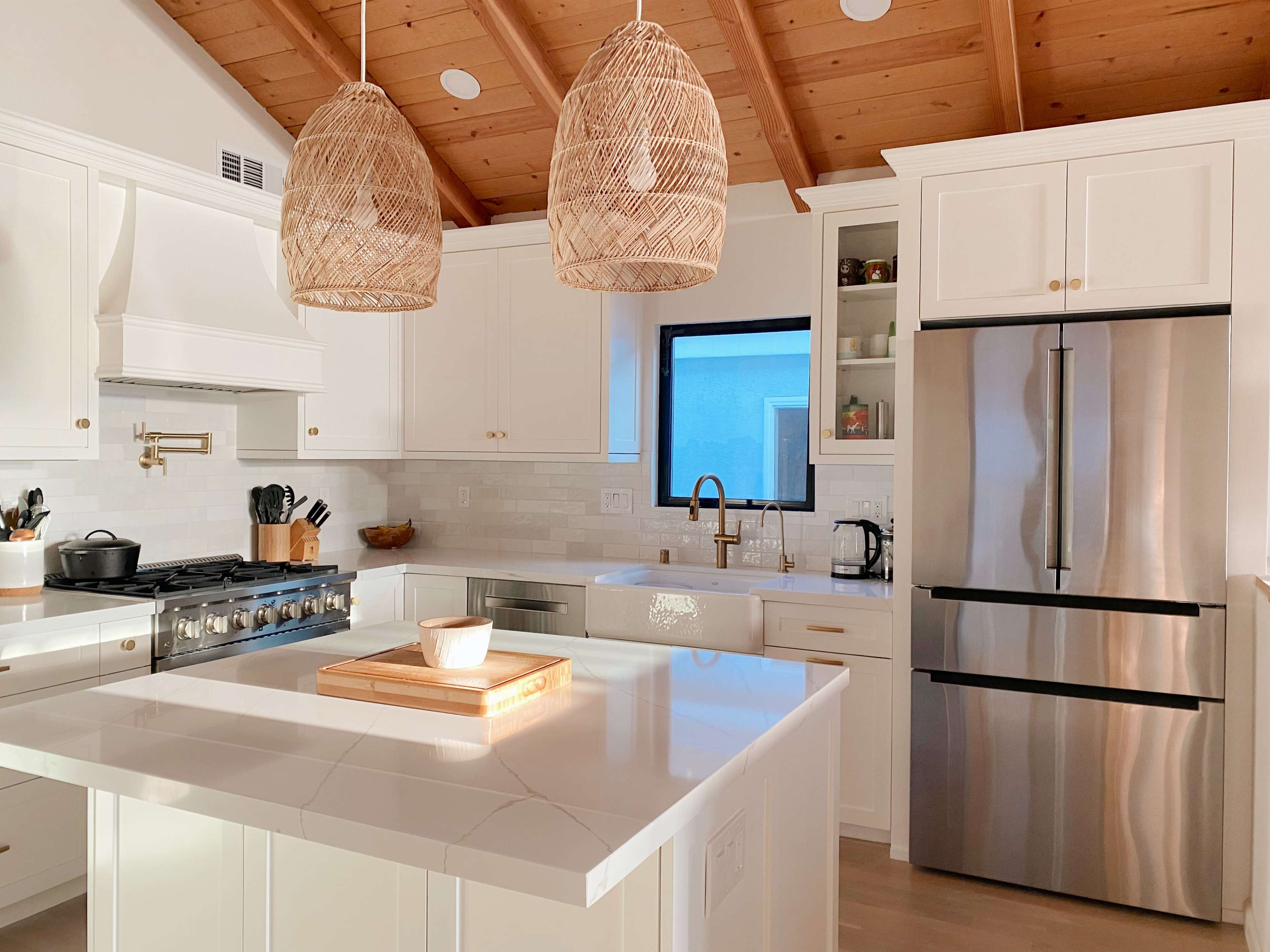 A modern kitchen features white cabinetry, a large island with a wooden cutting board, and two woven pendant lights hanging above.