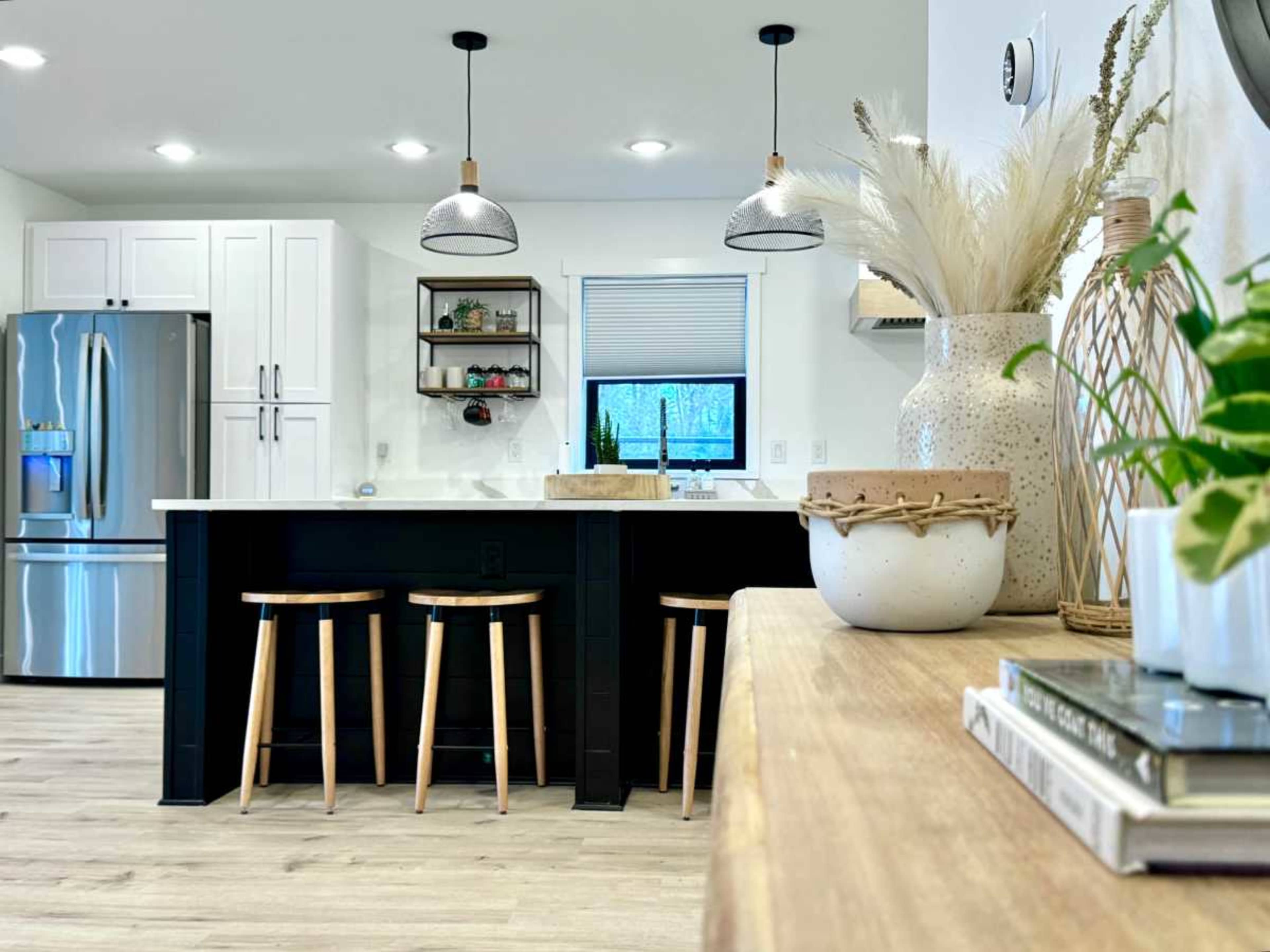 A modern kitchen features a central island with three wooden stools, stainless steel appliances, and decorative elements on a countertop.
