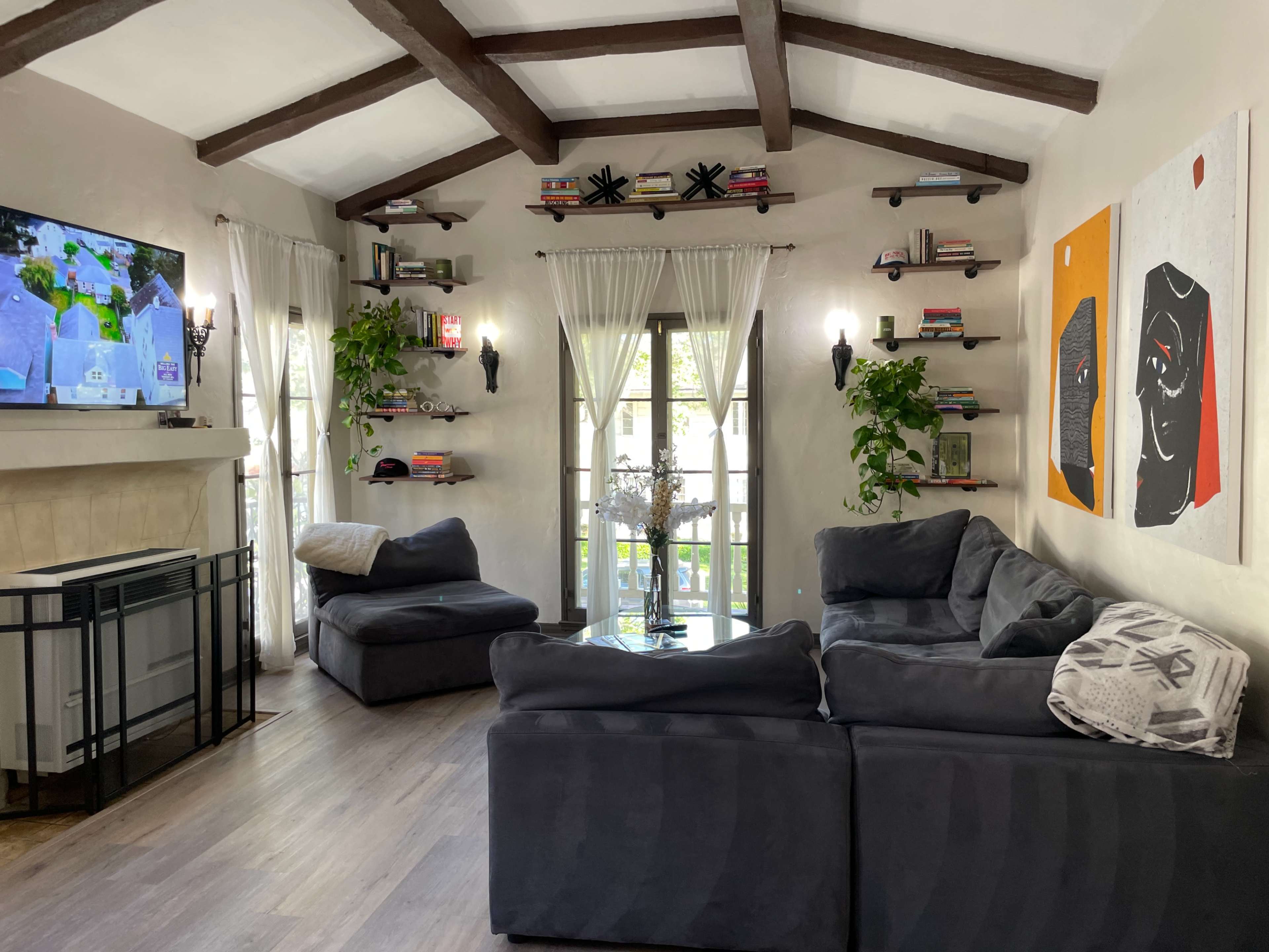 The image shows a cozy living room featuring a large sofa, a television mounted on the wall, and bookshelves with plants and decorative items.