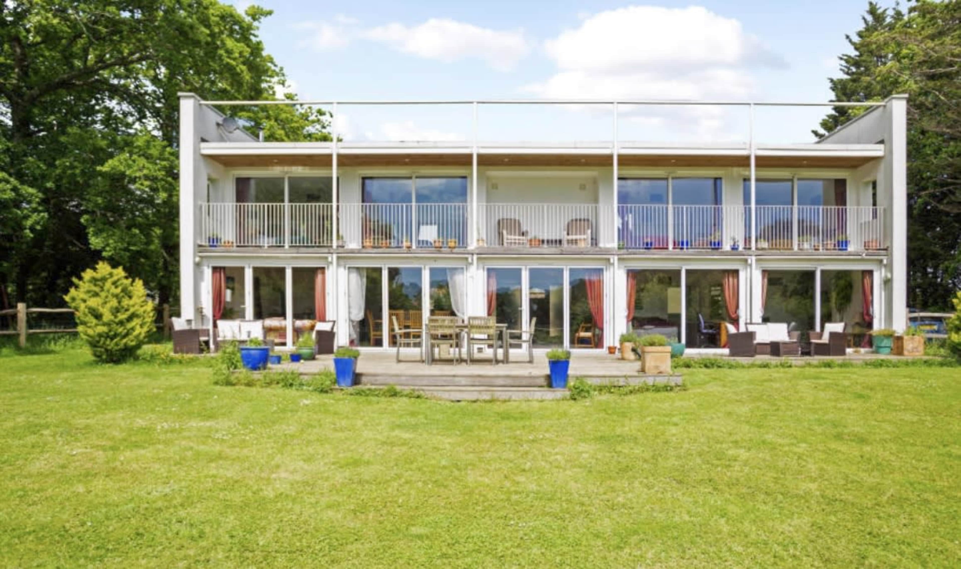 A modern two-story house with large glass doors and balconies overlooks a grassy yard.