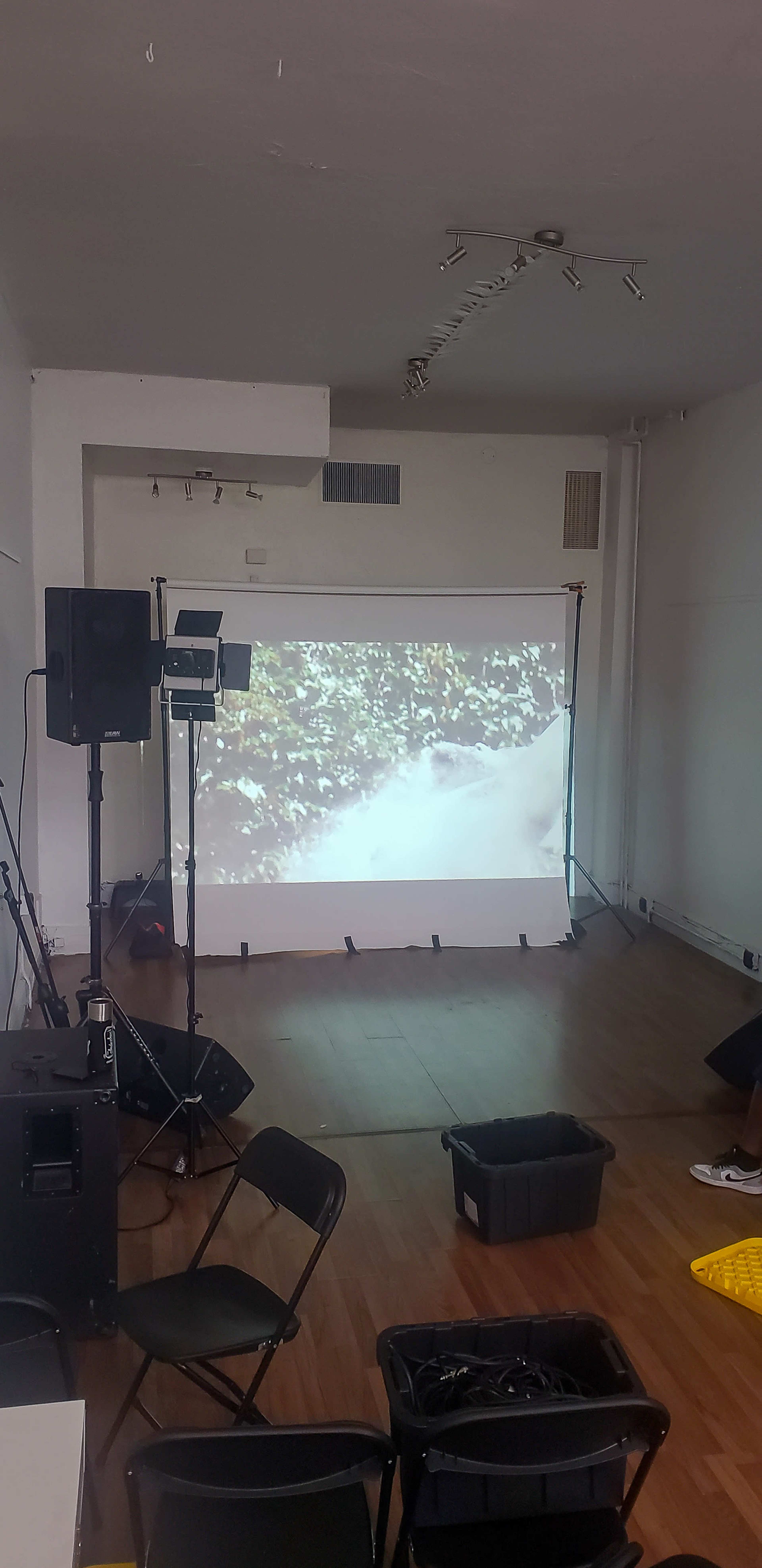 A projection screen displaying a video is set up in a sparsely furnished room, with sound equipment and a bucket on the floor.