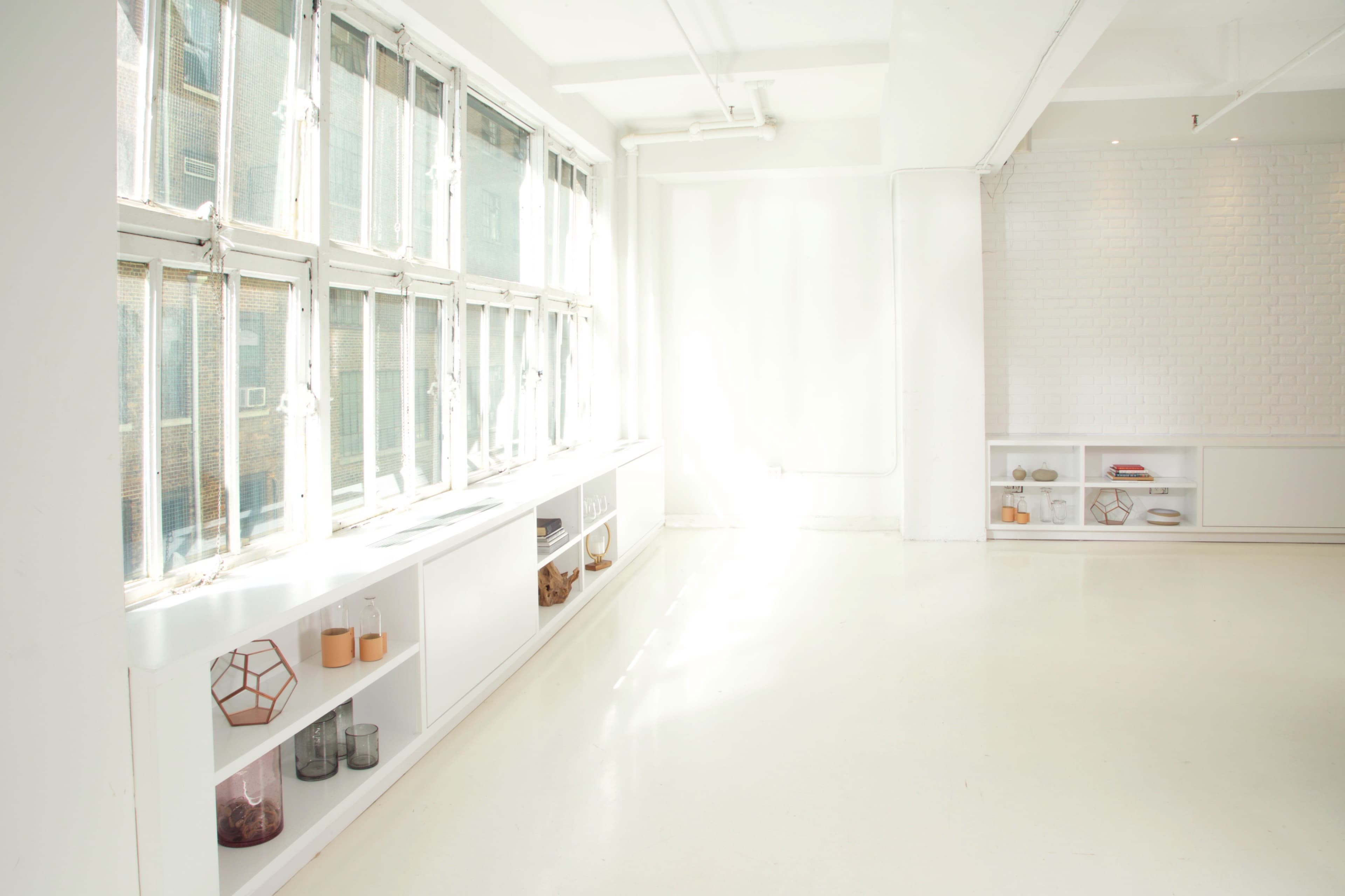 A bright, minimalist interior with large windows, white walls, and a sleek shelving unit along one side.