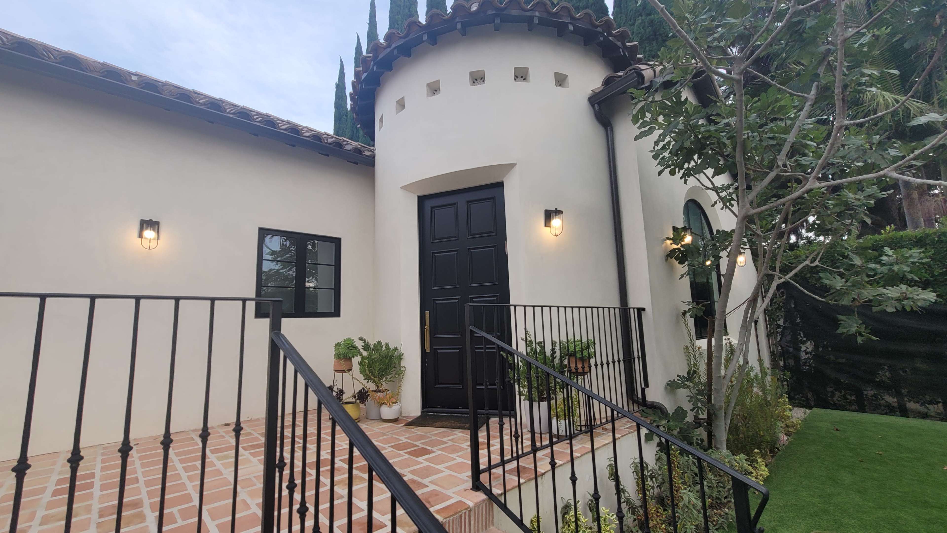A modern home features a round turret entry with a black front door, flanked by potted plants and wrought iron railings leading up to a tiled porch.